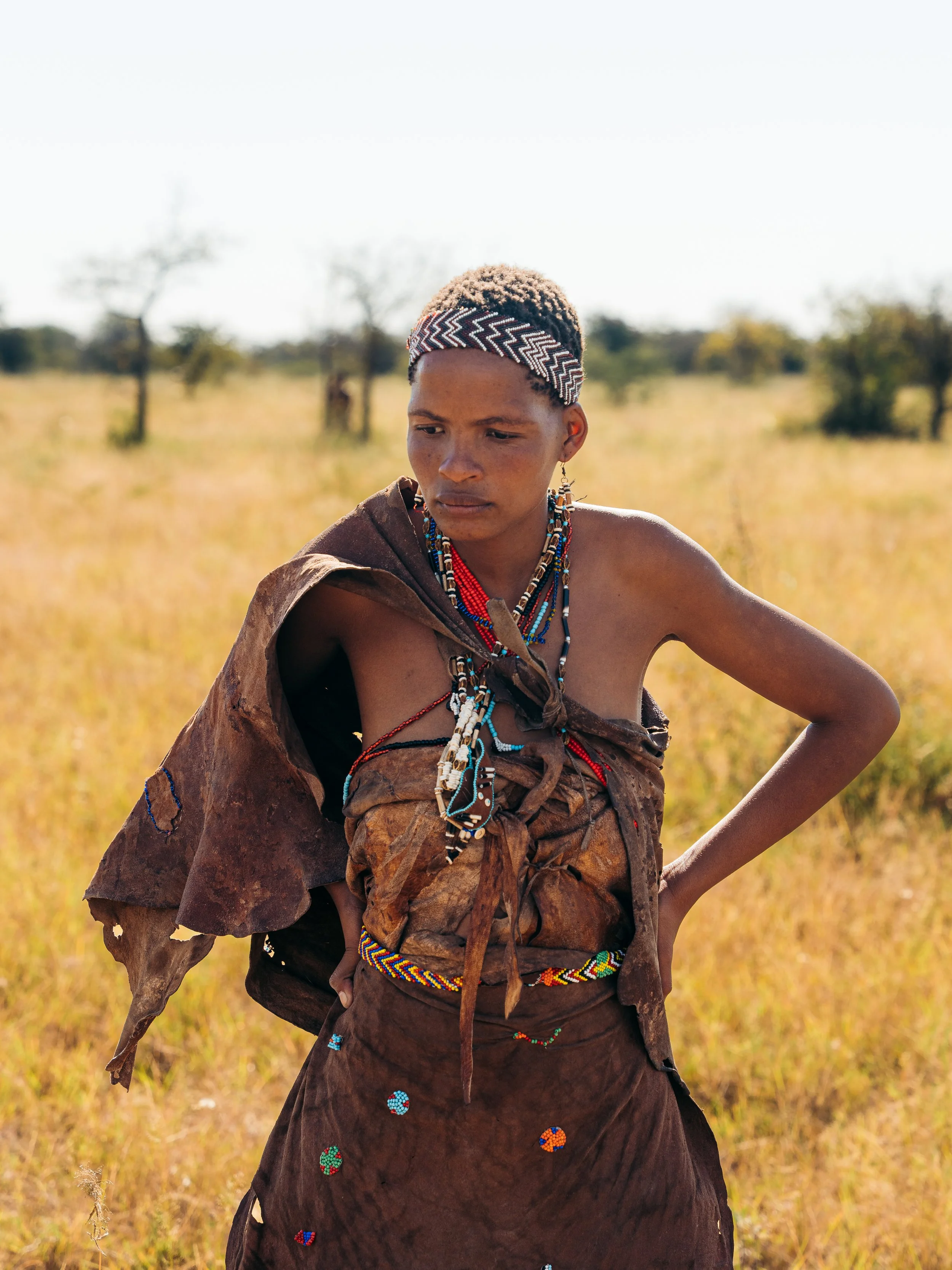 A young woman dressed in traditional Maasai attire, wearing beaded jewelry and a headband, standing outdoors in a grassy field with sparse trees in the background.