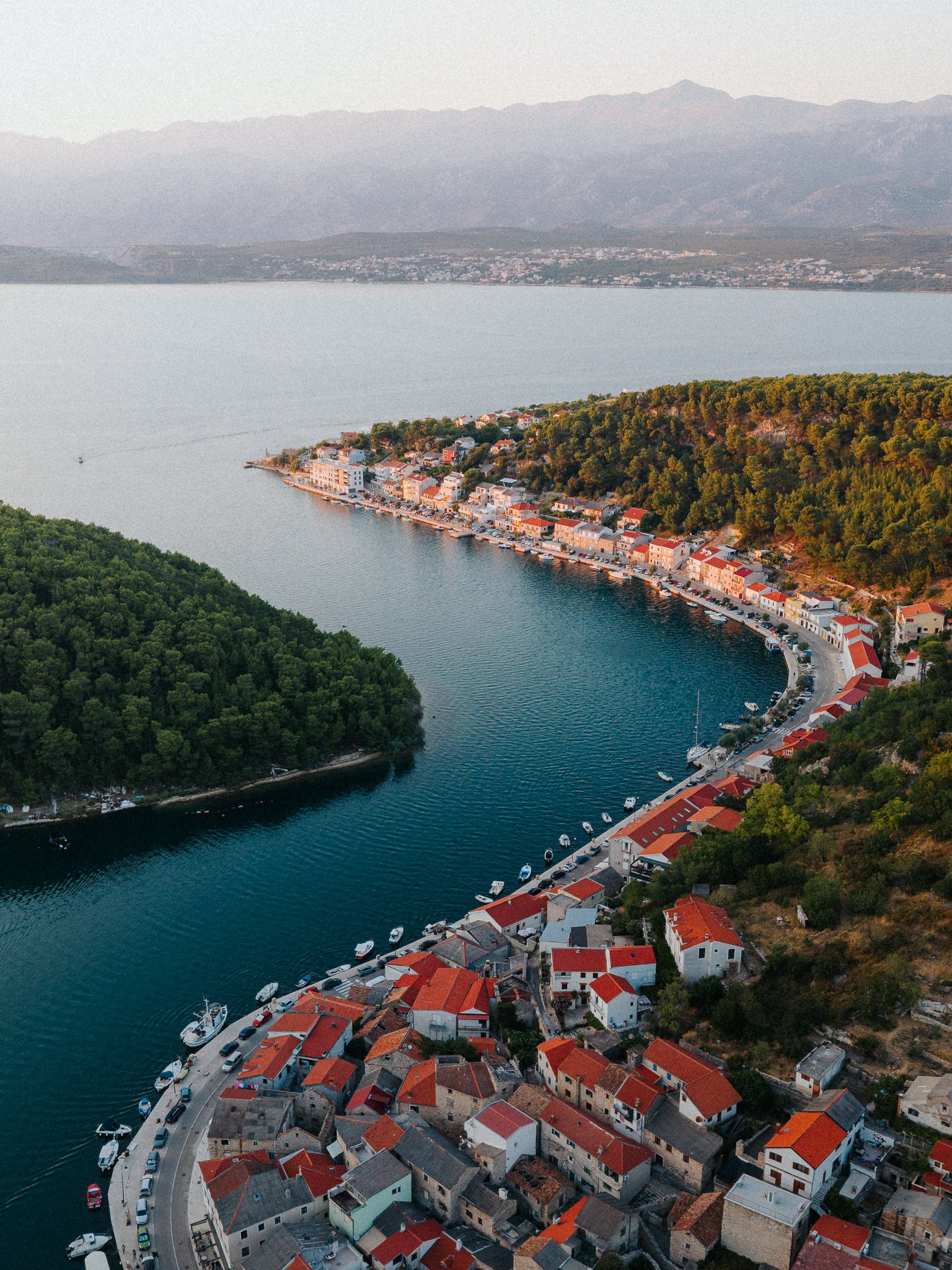 Aerial view of a coastal Mediterranean town with red-tiled roofs, houses along a waterway, surrounded by green hills and a large body of water with distant mountains.