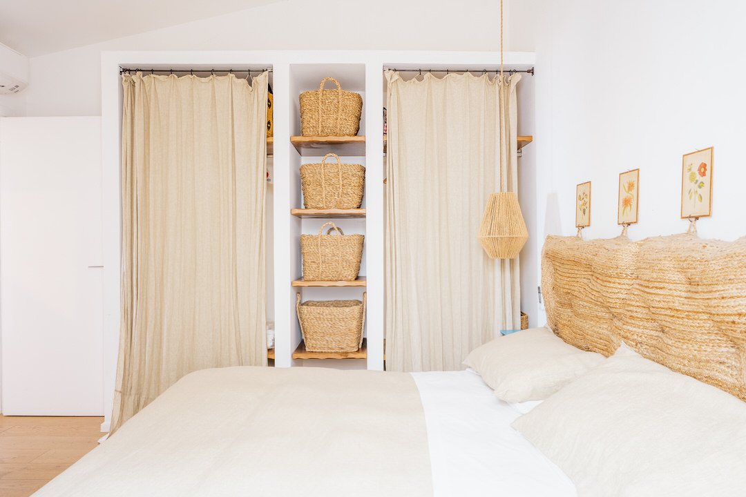 A bedroom with a bed, woven headboard, beige pillows, and white bedding, featuring a wall with hanging art and a shelving unit with wicker baskets behind beige curtains.