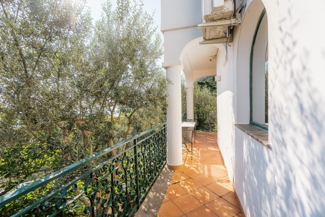 A balcony with orange tiled floor, white walls, and a decorative green metal railing, overlooking trees with sunlight filtering through.