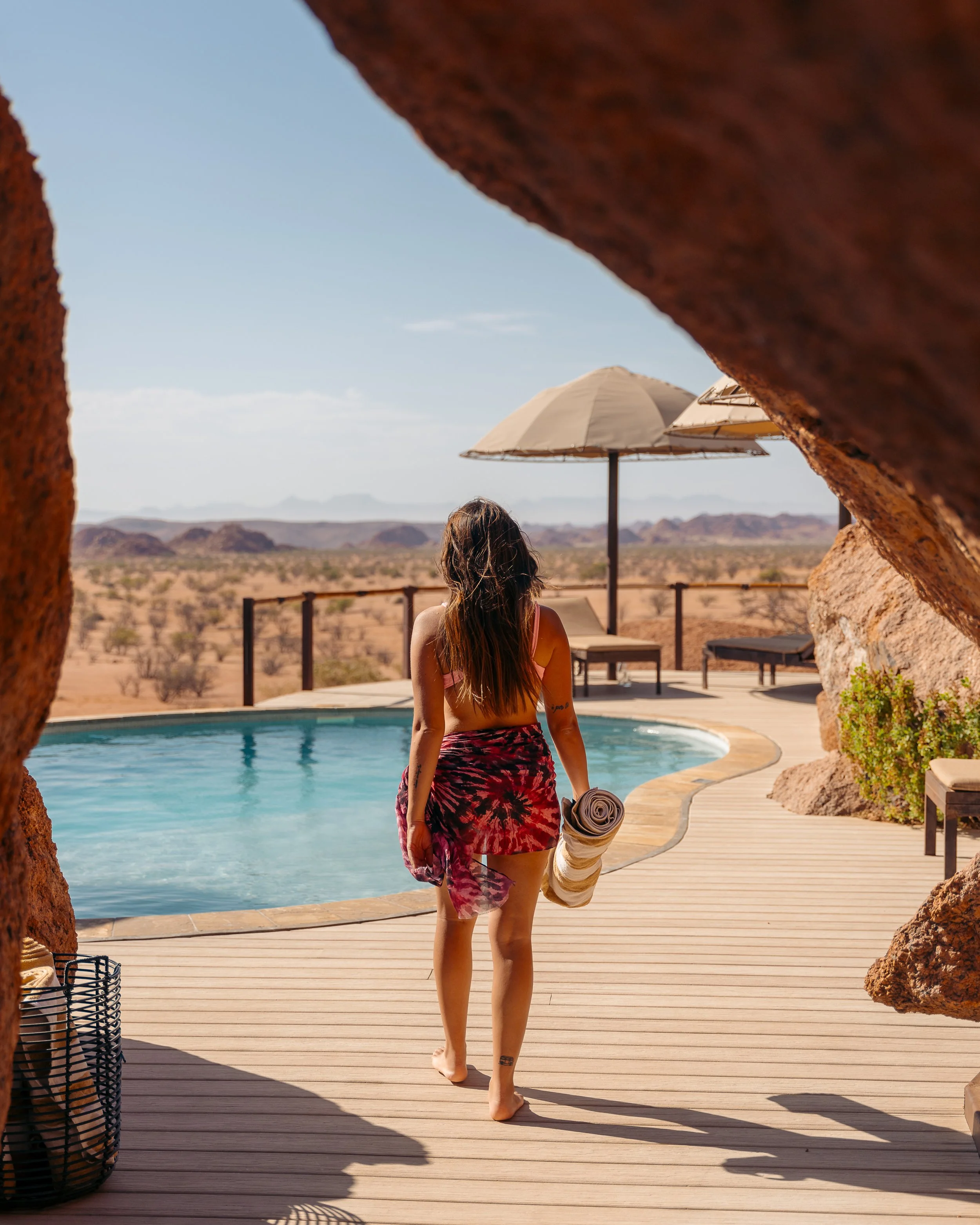 A woman with long brown hair, wearing a bikini top and a tie-dye skirt, walks barefoot on a wooden deck toward a pool in a desert landscape. She carries a rolled towel in her right hand. The scene is framed by large reddish rocks with an umbrella and