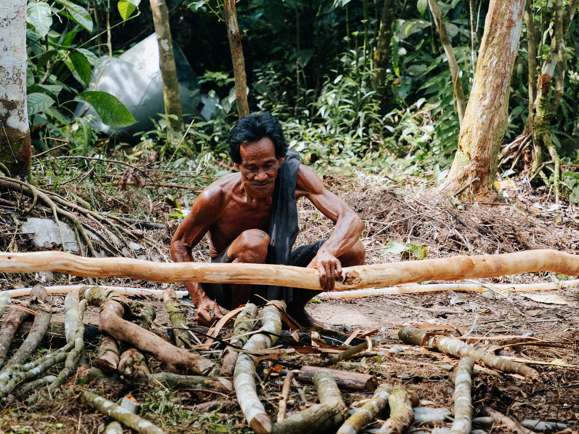 An elderly man with dark skin, black hair, and a mustache, shirtless, kneeling in a rainforest, cutting a long wooden stick with a machete.