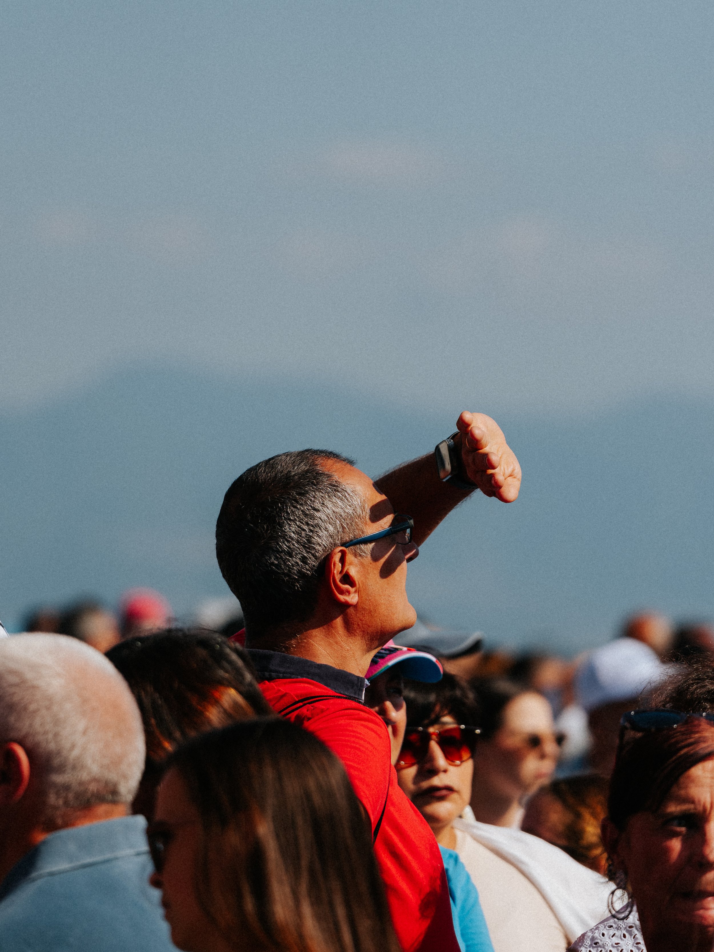A man with short gray hair wearing glasses, a red jacket, and a smartwatch is saluting in a crowd during daylight. The crowd includes diverse individuals, some women wearing glasses and hats, under a clear sky.