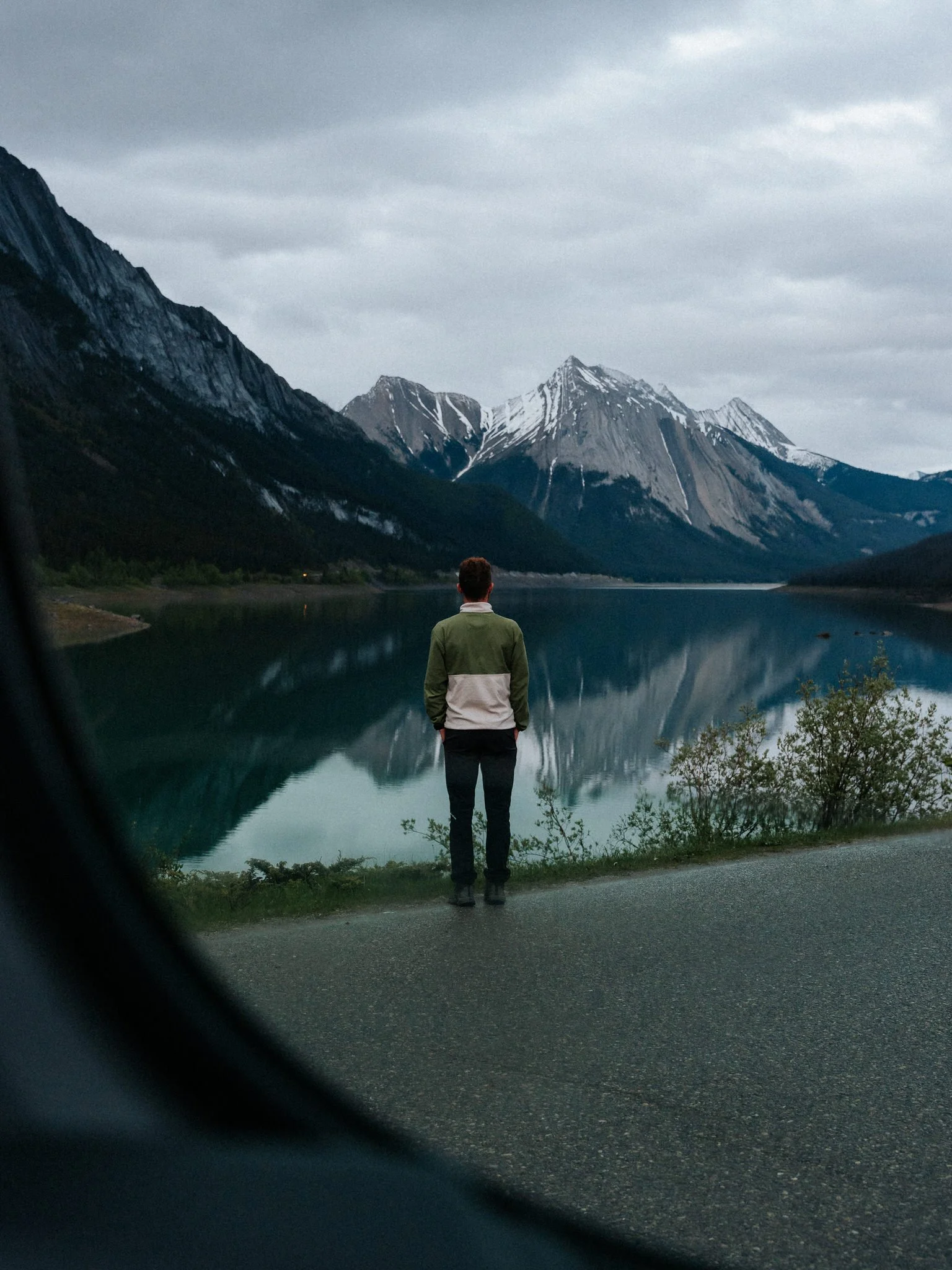 A person stands by a reflective mountain lake facing snow-capped mountains under a cloudy sky.
