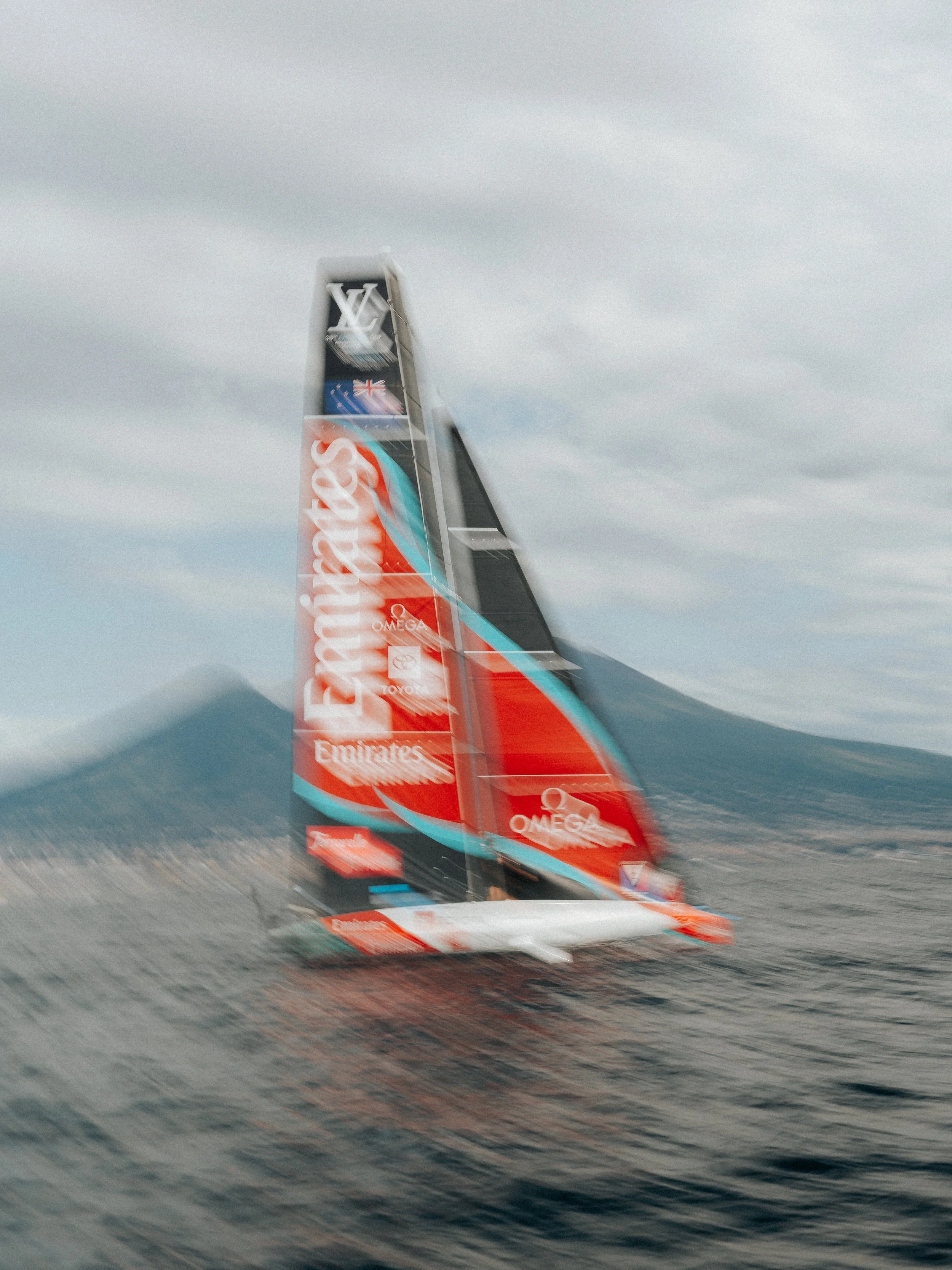 A sailboat racing on the water with advertisements on its sail, including brands like Emirates, Omega, and Renault, under cloudy skies.