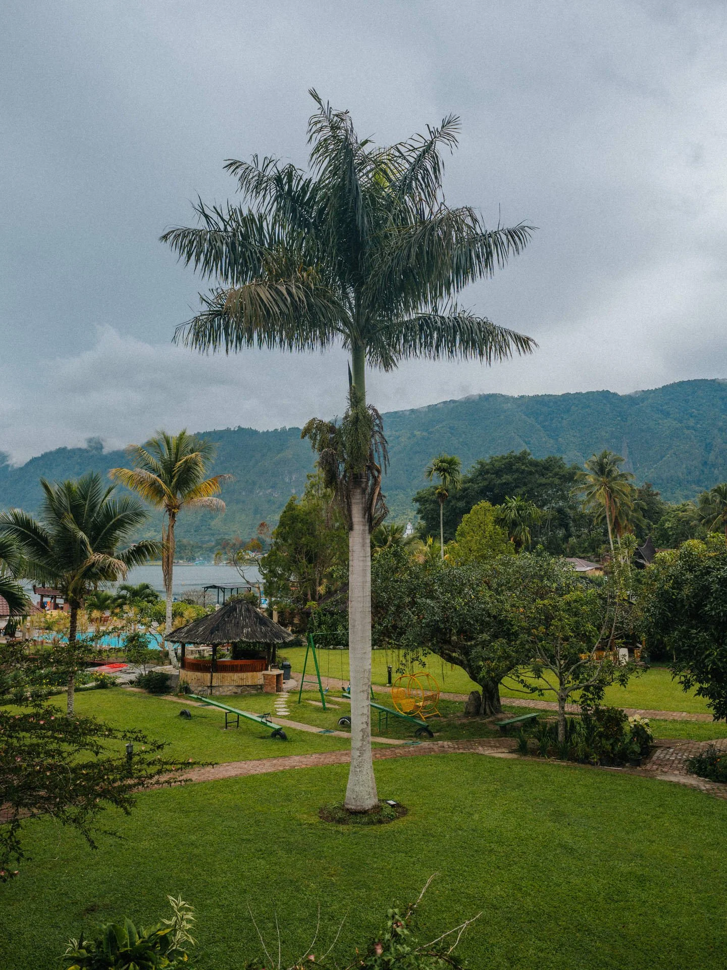 A park with lush green grass, palm trees, a small thatched-roof hut, a picnic table, and playground equipment against a backdrop of hills and a cloudy sky.