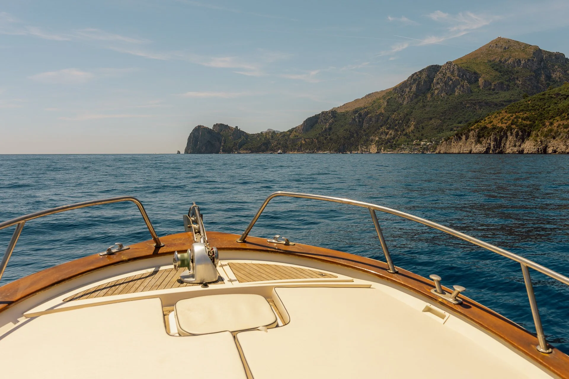 View from the deck of a boat showing calm blue sea, distant coastline with green hills and mountains under a partly cloudy sky.