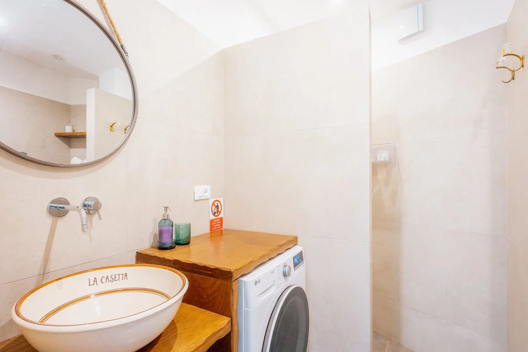 Bathroom with a wooden countertop holding soap and three bottles, a washing machine, a boat-shaped sink with "LA CASETTA" inscribed, a round mirror, and a shower area in the background.