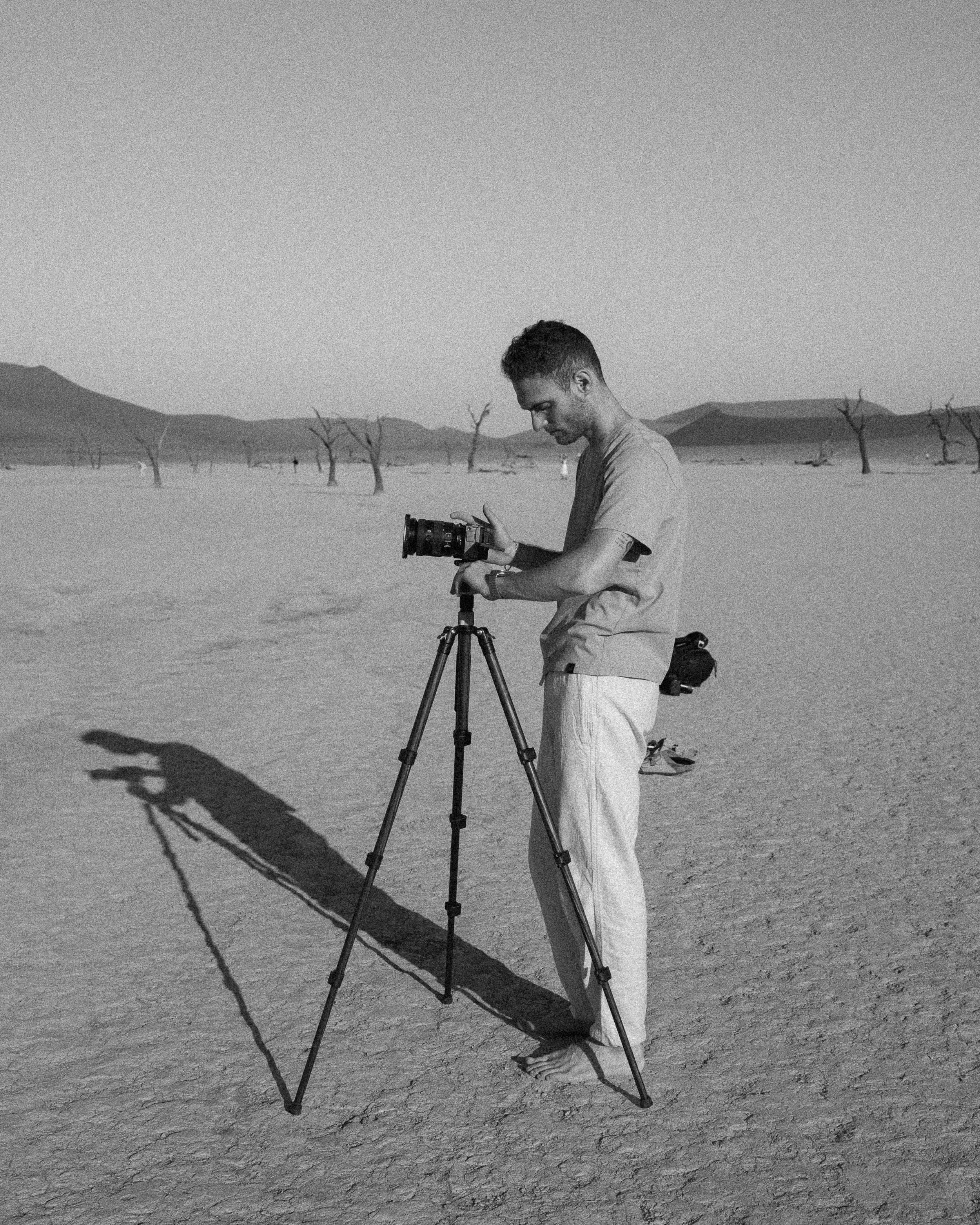 A man setting up a camera on a tripod in a dry, desert landscape with sparse, leafless trees and distant mountains.