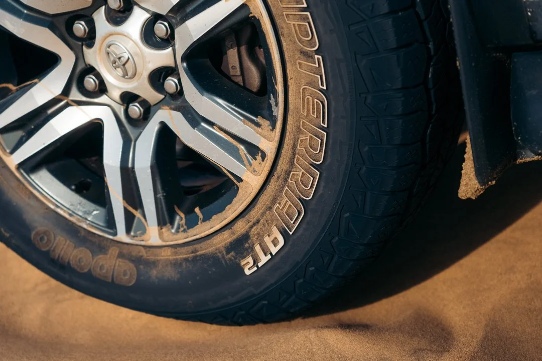 Close-up of a Toyota vehicle tire with dirt on the sidewall, mounted on a black alloy wheel with a silver finish and six spokes.
