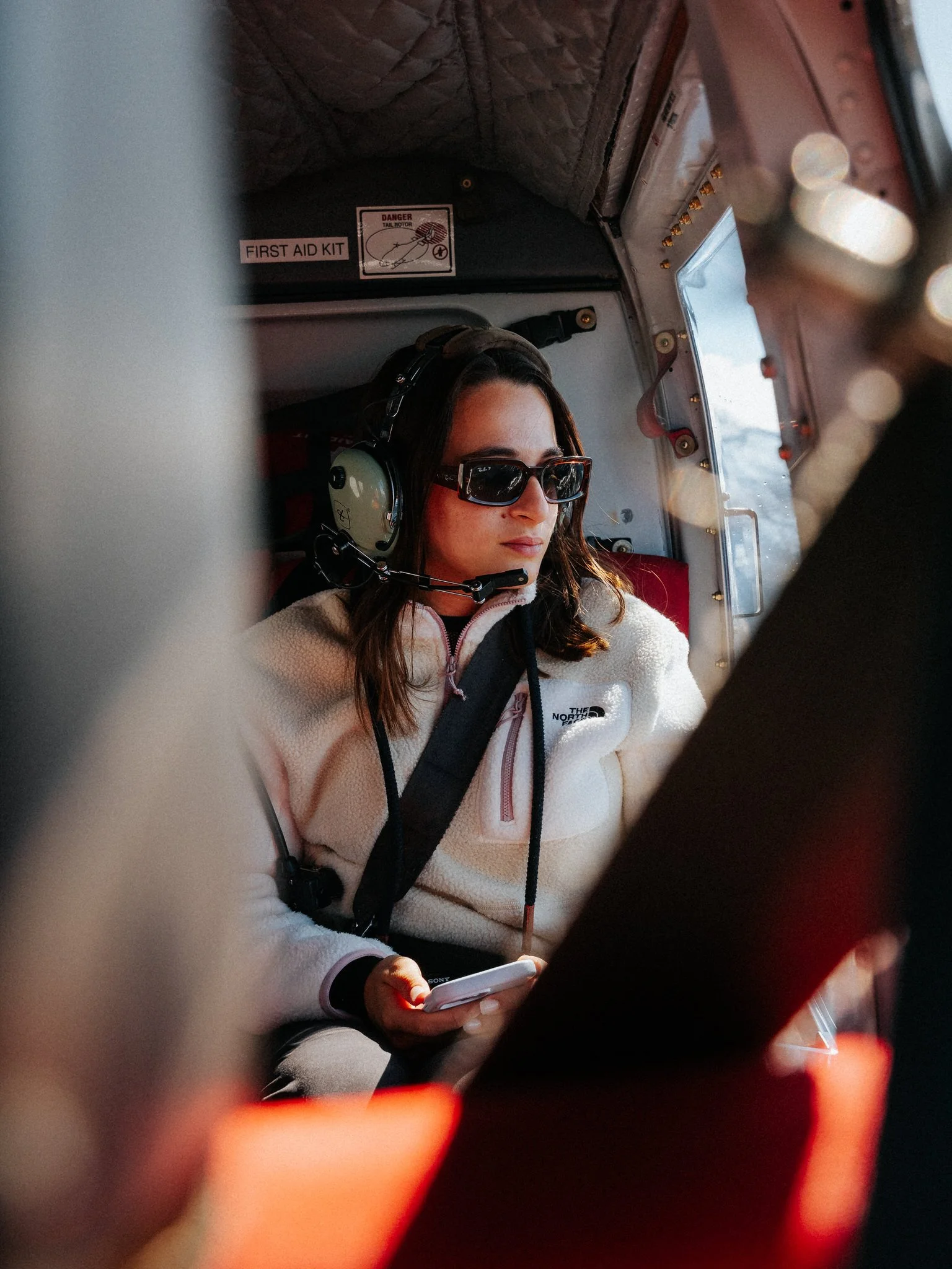 A woman sitting inside an aircraft wearing sunglasses and a headset, holding a smartphone, with first aid kit and aircraft interior visible.