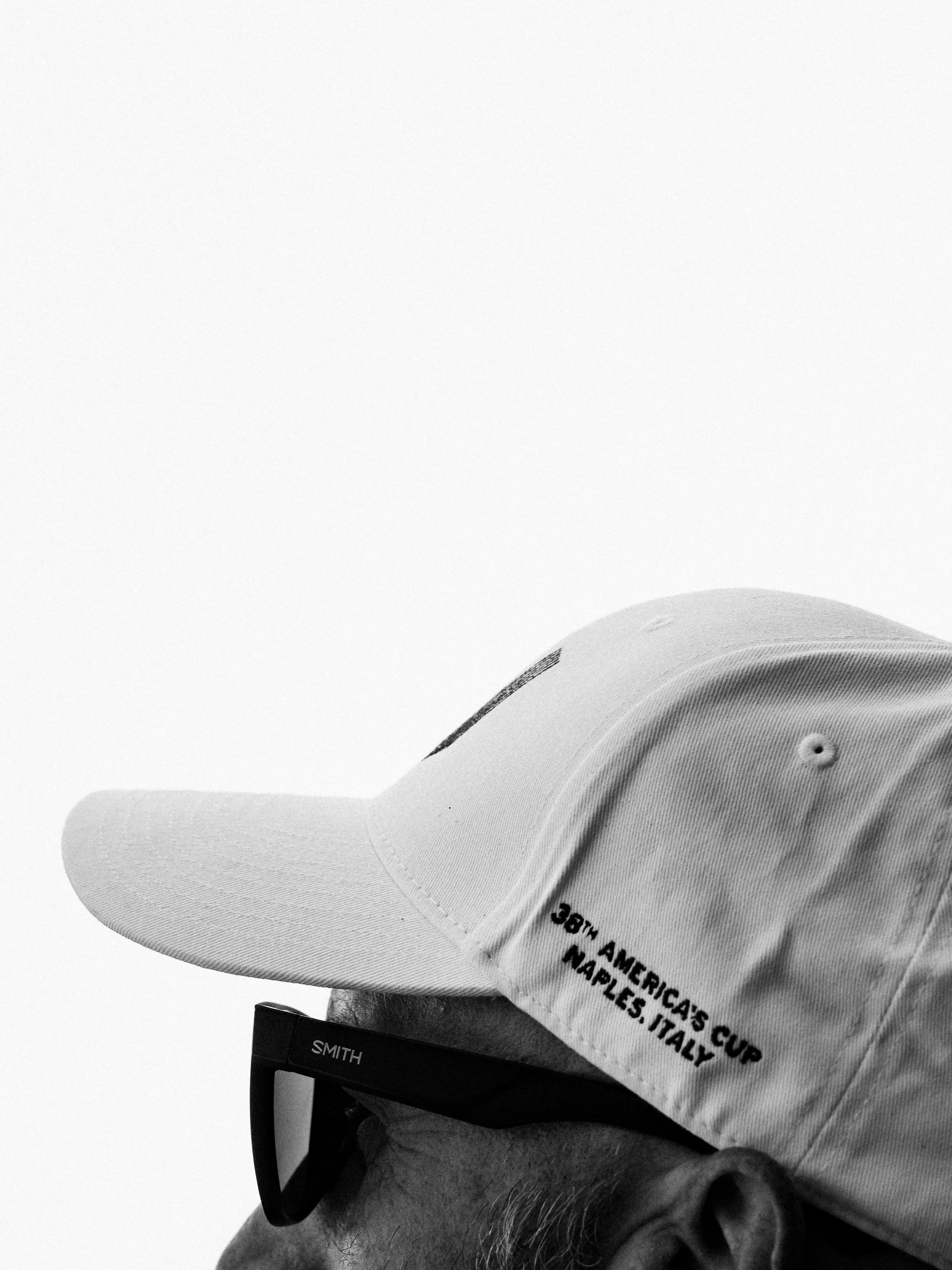 A black and white photo of a man wearing glasses and a white baseball cap with text. The cap reads 'Made in America, Cup Naples, Italy'.