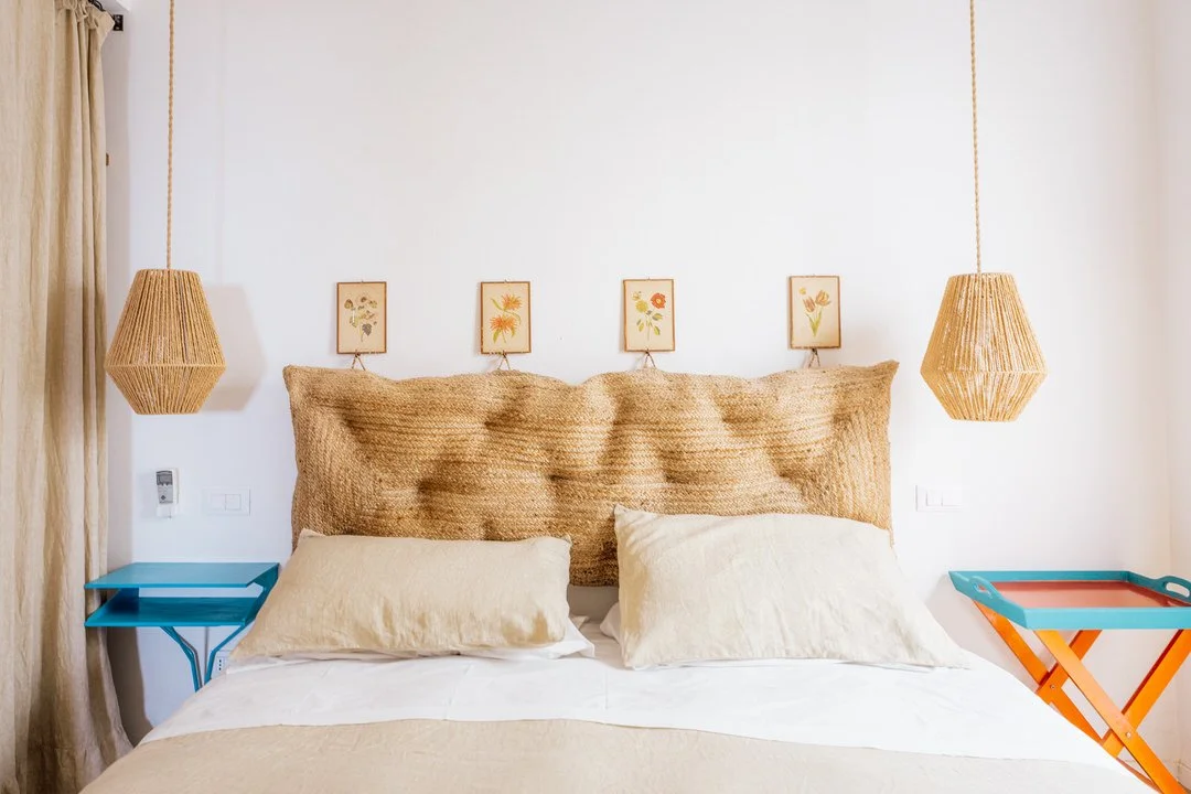 A bedroom with a large headboard, two pillows, hanging wicker lamps, and small colorful bedside tables.
