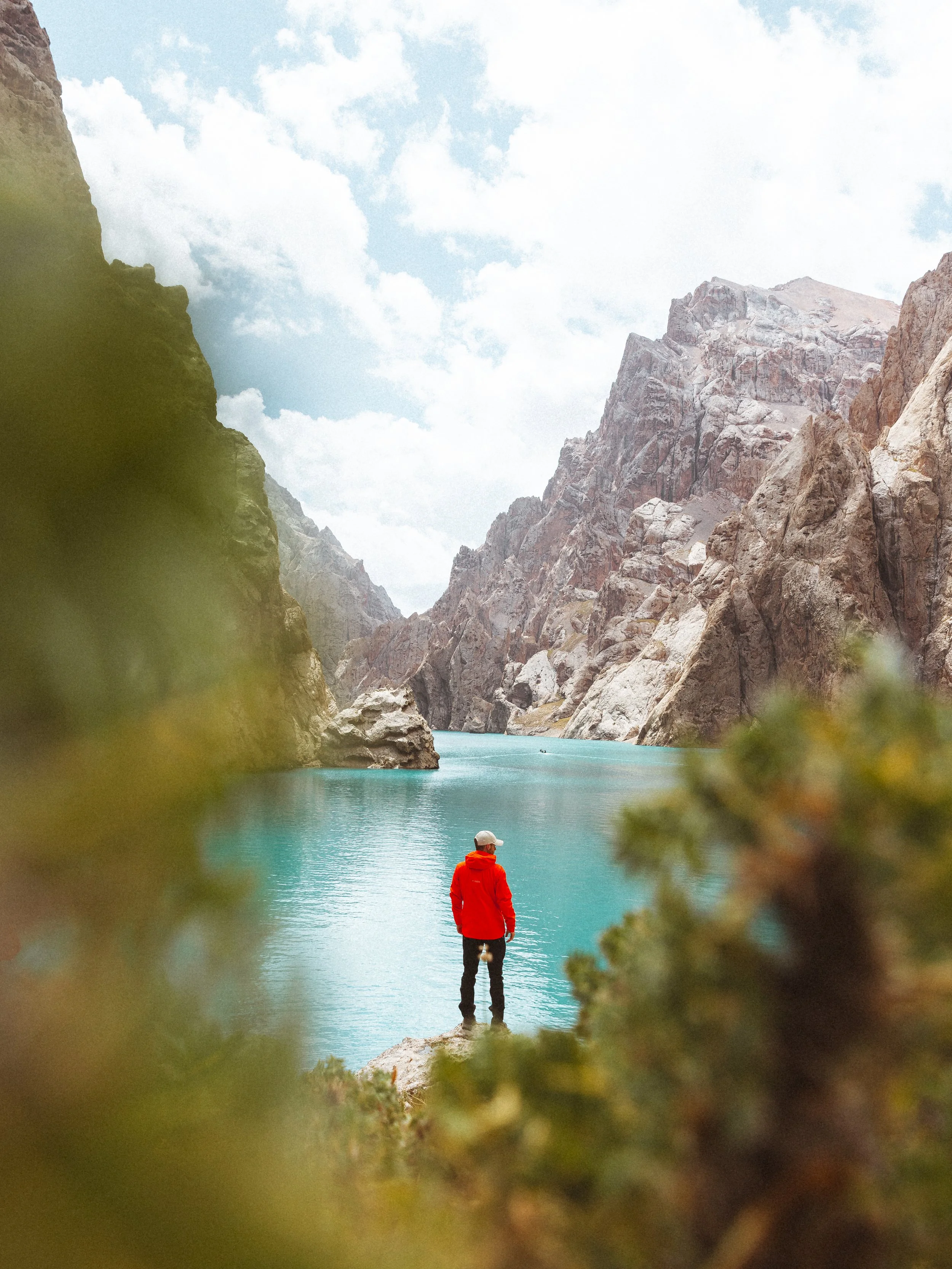A person in a red jacket and white hat stands on a rocky outcrop overlooking a turquoise mountain lake, surrounded by steep rocky cliffs and partly cloudy sky.