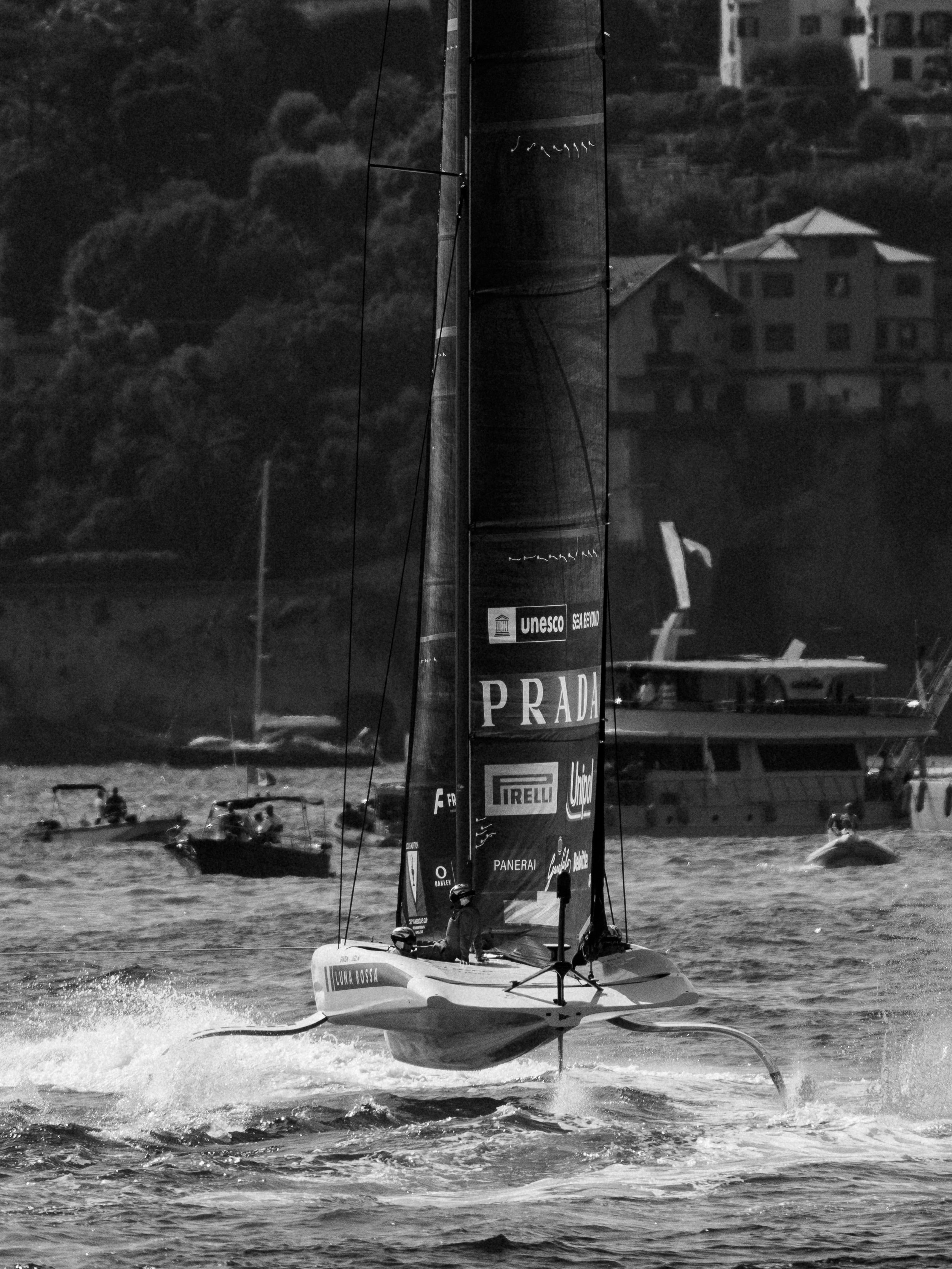A sailboat racing on the water, with a tall sail covered in logos and text, including 'Prada' and 'UNESCO.' Several smaller boats and people are visible in the background, with buildings on a hill and trees behind.