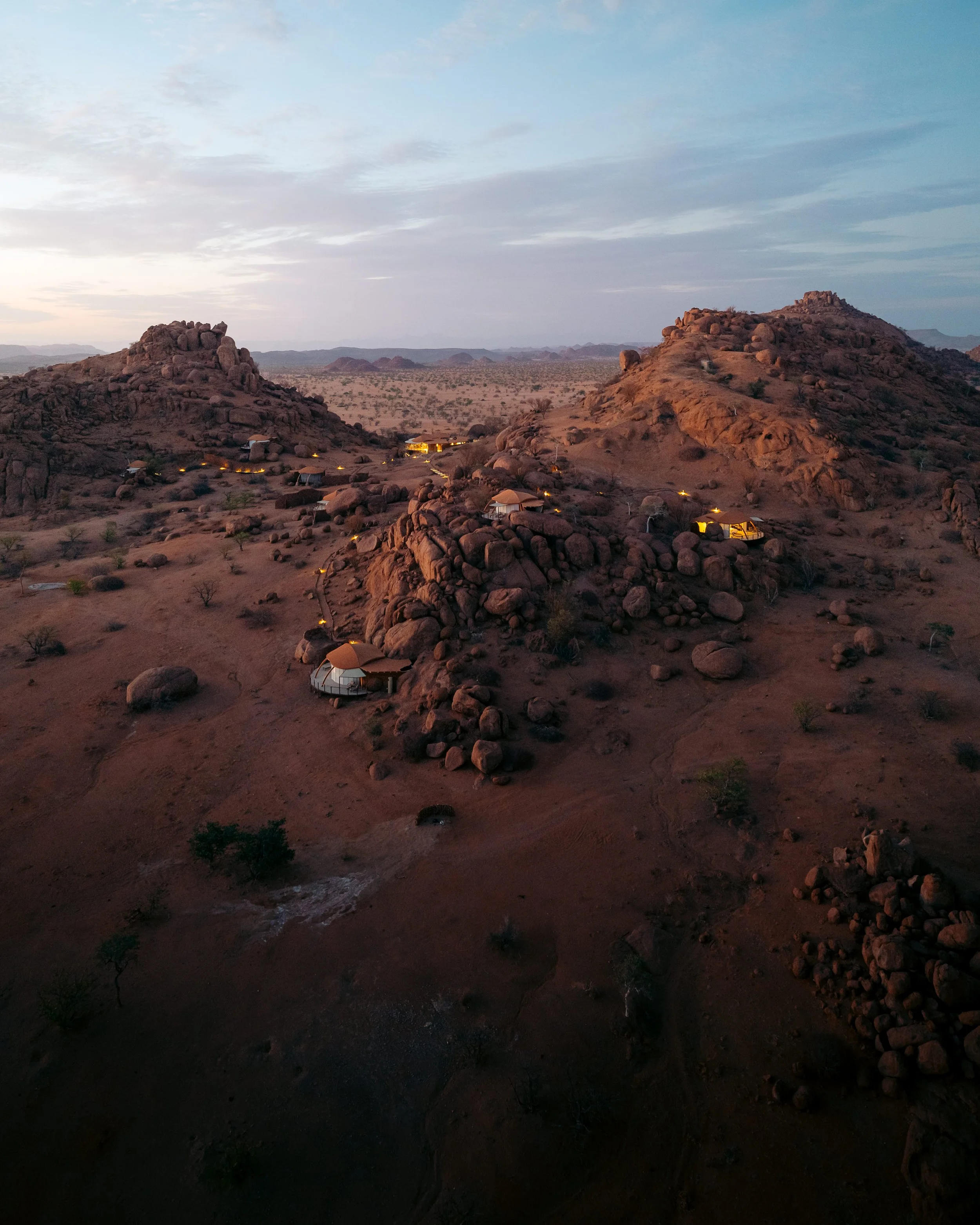 Aerial view of a desert landscape with rocky hills and small illuminated tents or structures scattered across the terrain at dusk.