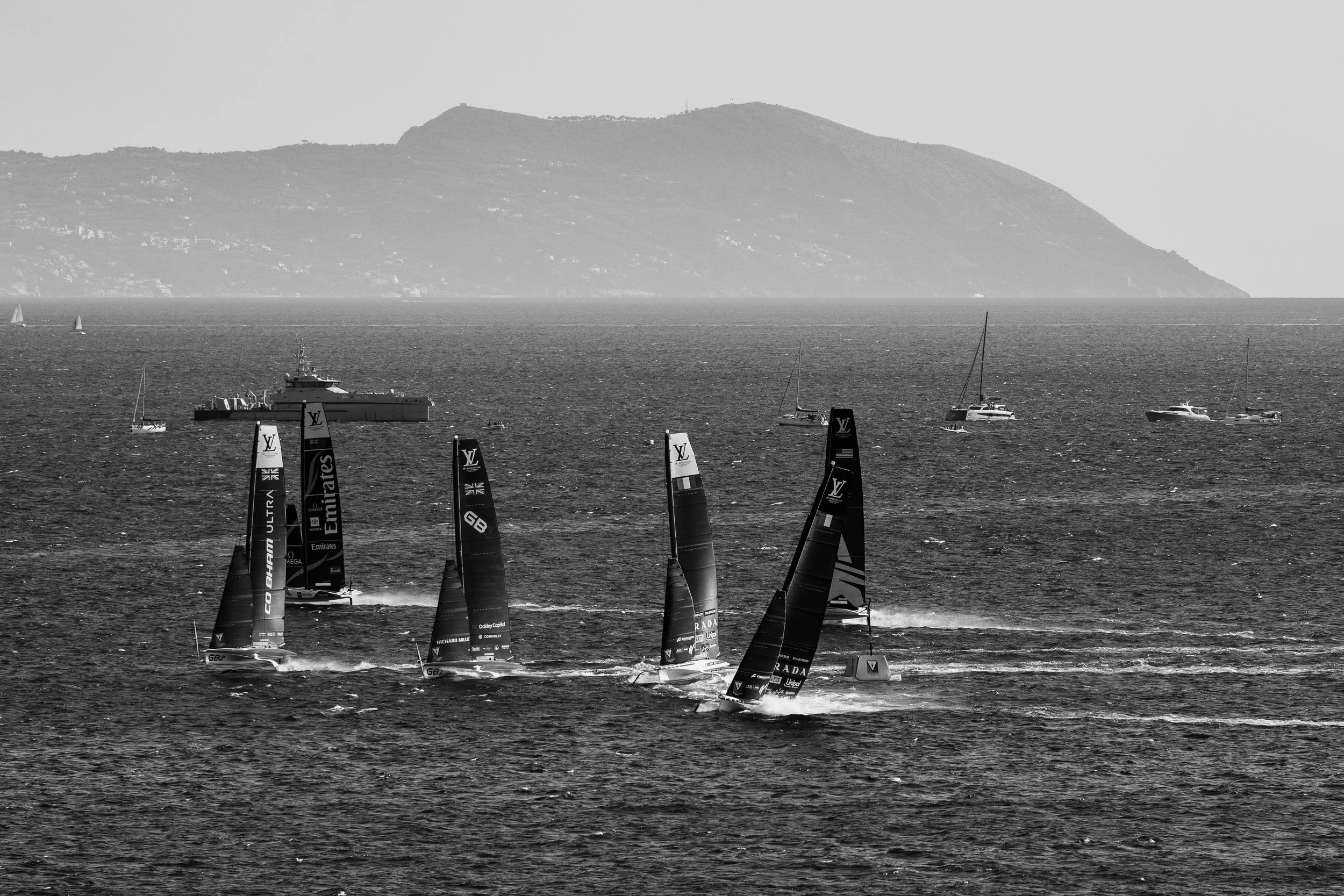 Black and white photo of sailboats racing on the water, with a mountainous coastline in the background.
