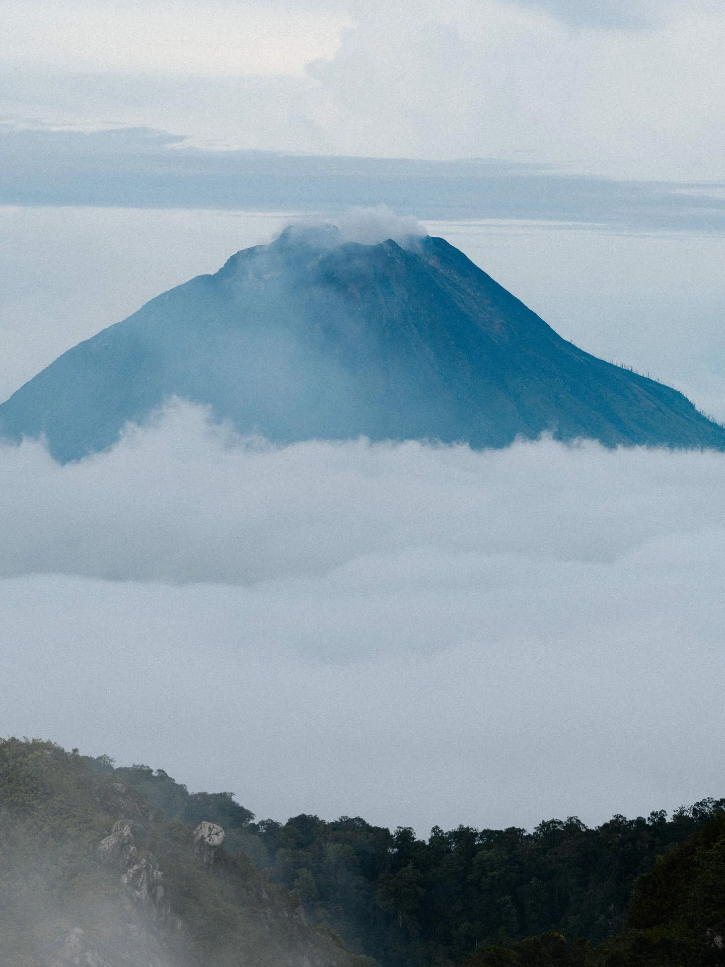 A volcano surrounded by clouds, with a forested landscape in the foreground.
