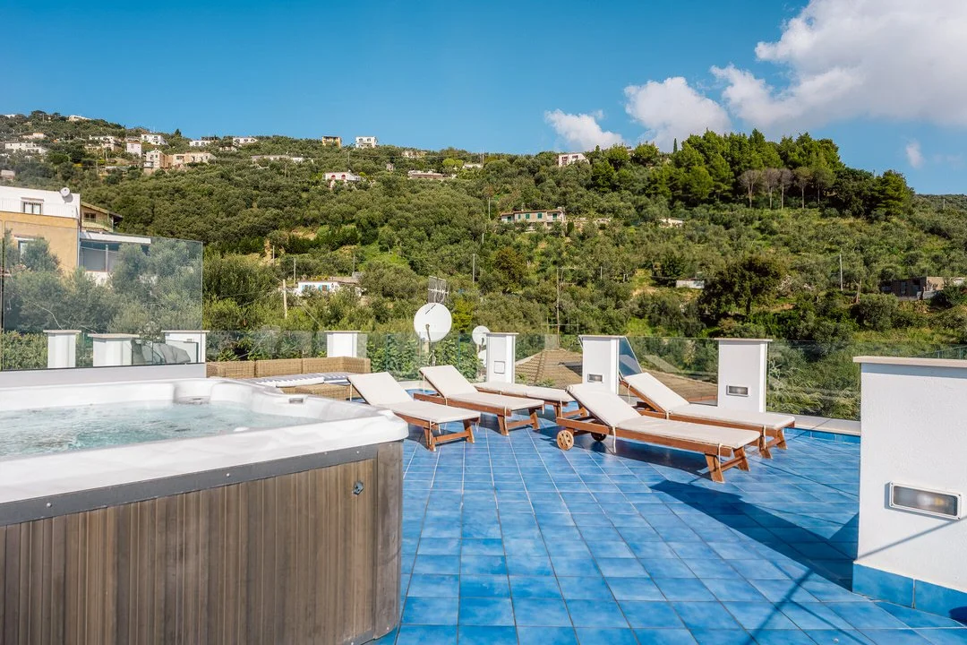 Rooftop terrace with four lounge chairs, a hot tub, and a view of green hills and houses, under a partly cloudy sky.