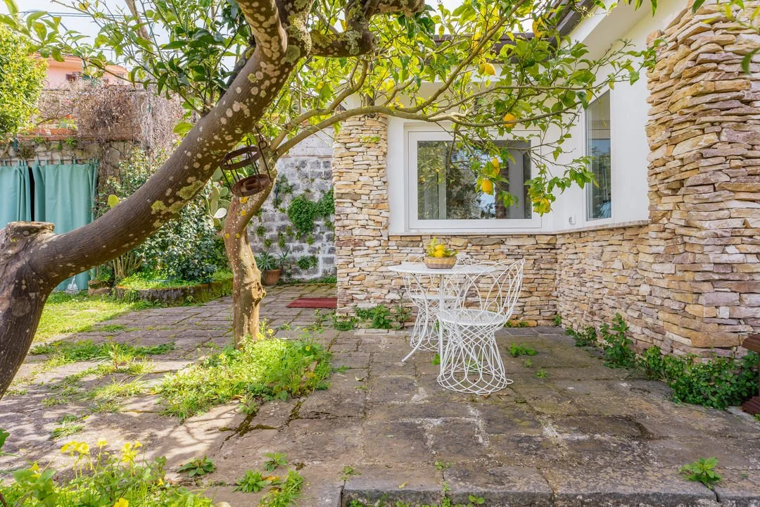 A cozy outdoor patio area with stone walls, a large tree, a small white wrought iron table with matching chairs, and green foliage.
