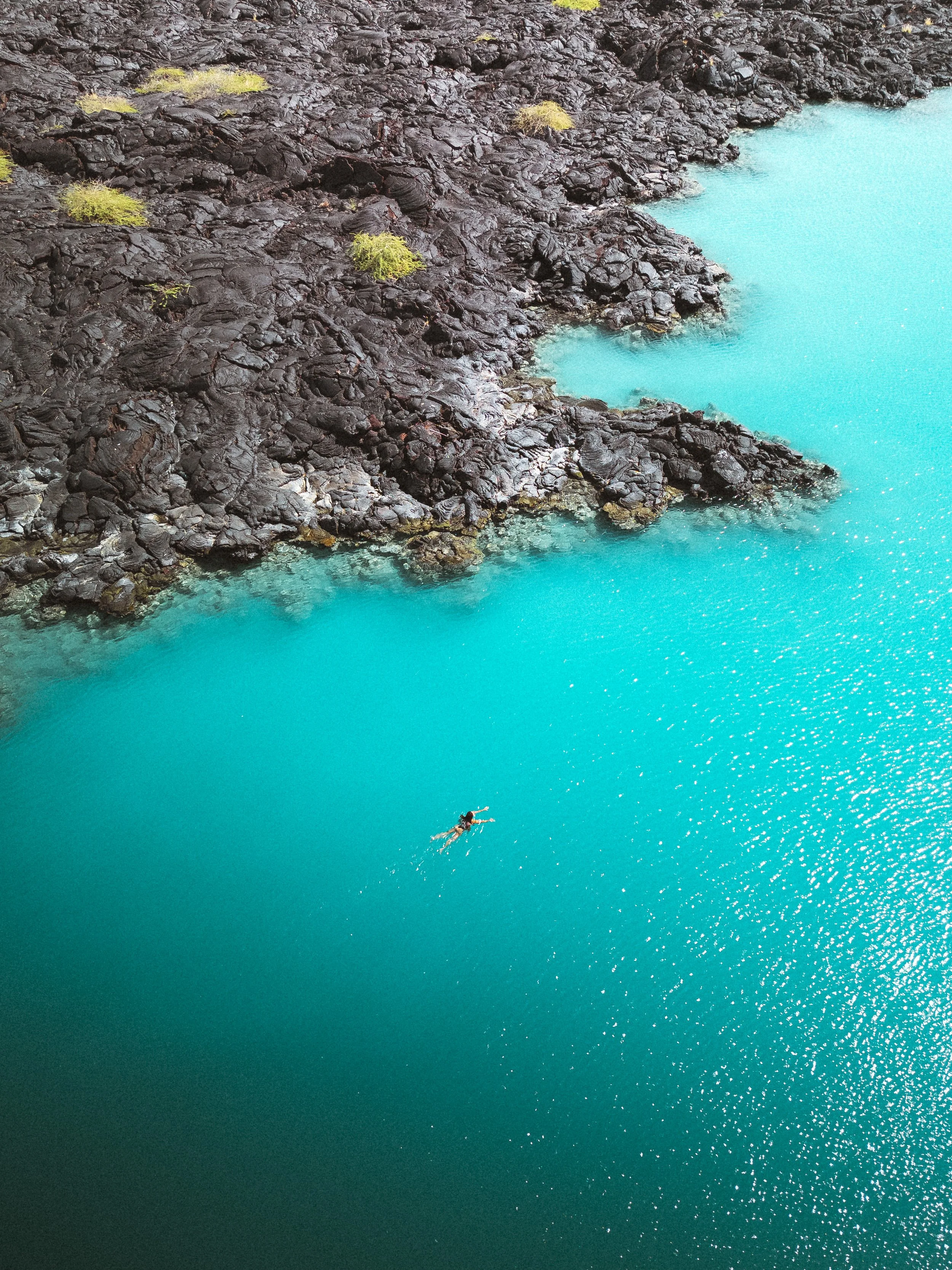 A person swimming in a turquoise lake near a rocky, dark shoreline with sparse green vegetation.