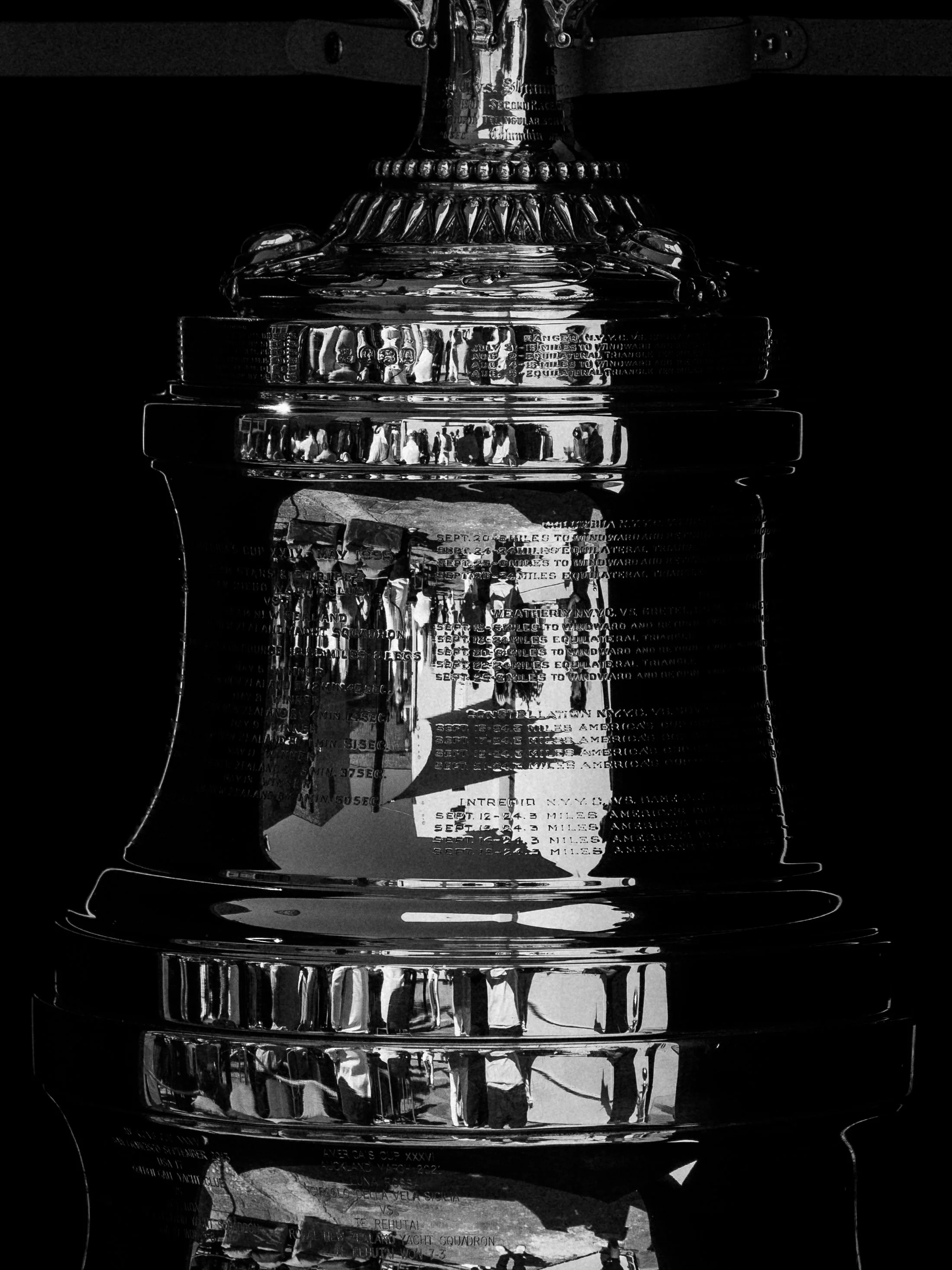 Black and white close-up of a shiny trophy with inscriptions and reflections, with people and surroundings visible in the reflections on the trophy's surface.