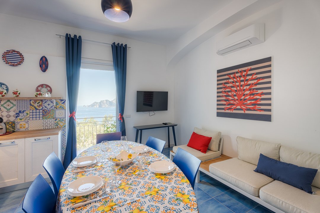 A dining area with a table set with plates, glasses, and a lemon bowl, next to a window with blue curtains showing a scenic seaside view, with colorful patterned tiles on a sideboard, a wall-mounted TV, a beige sofa with red and blue cushions, a mode