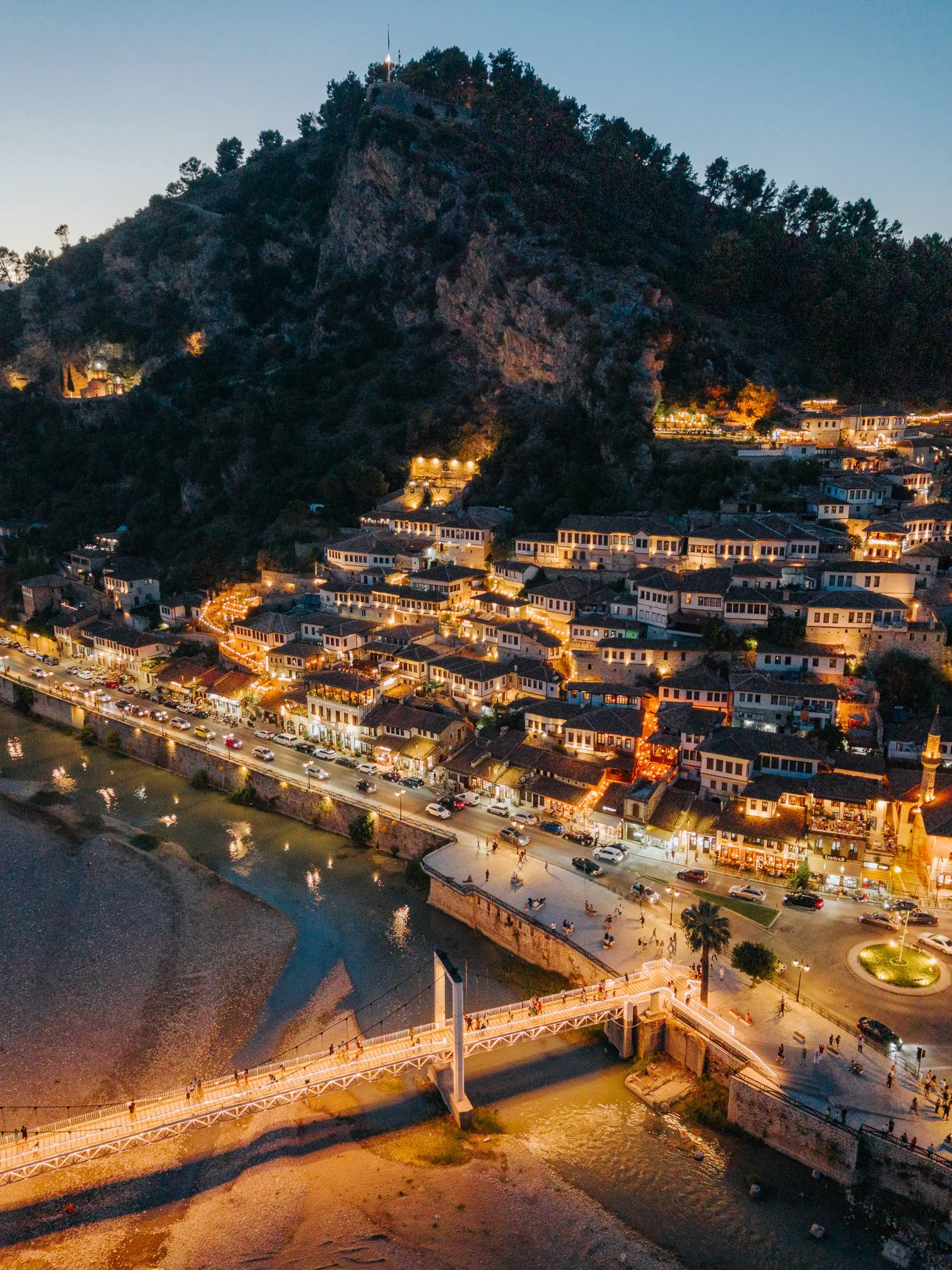 Aerial view of a hillside town at dusk with buildings and streets lit up, a river running through the town, and a small bridge crossing the river.