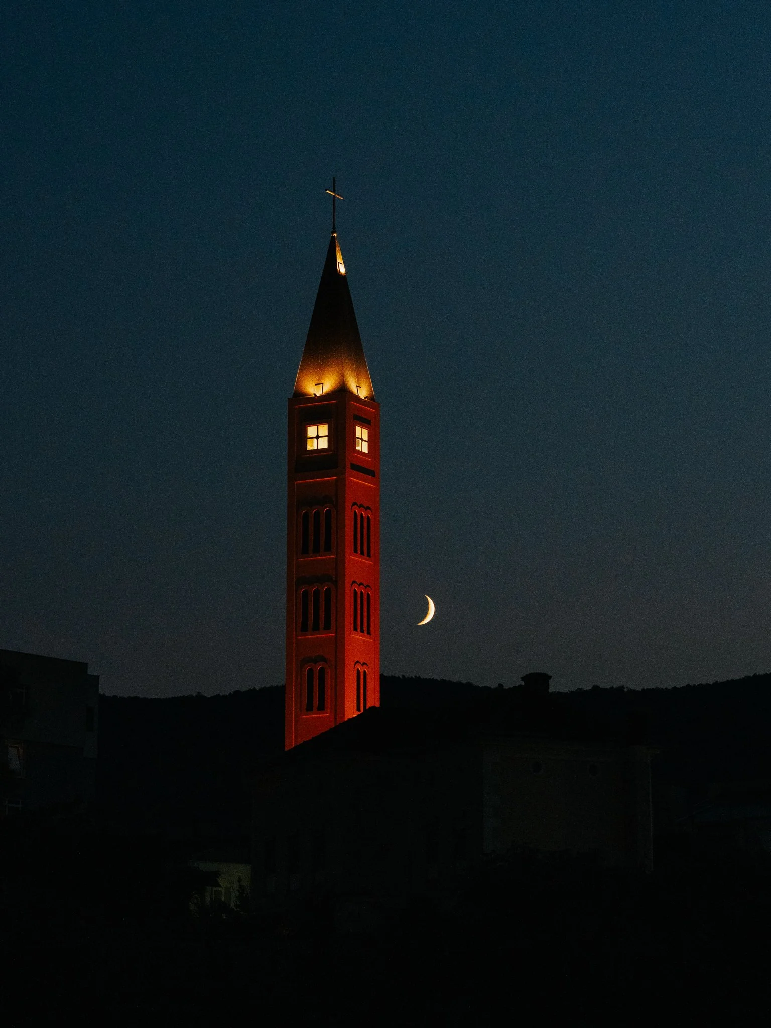 Night view of a lit red church steeple with a cross on top, with a crescent moon in the dark sky.