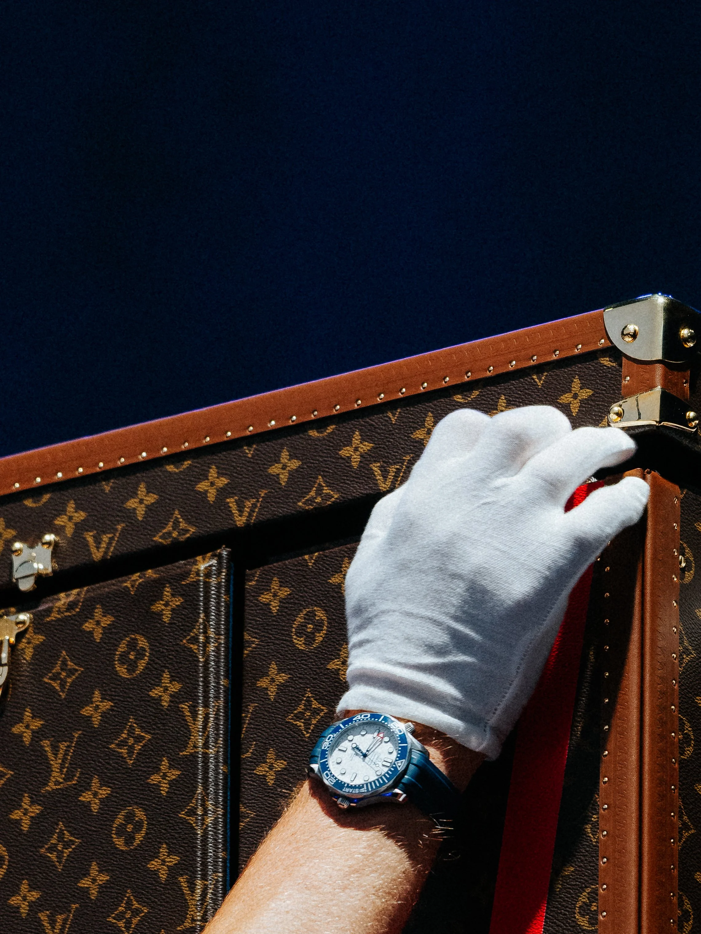 A person wearing a white glove holding a Louis Vuitton monogram patterned suitcase at night. The person is wearing a blue and silver wristwatch.
