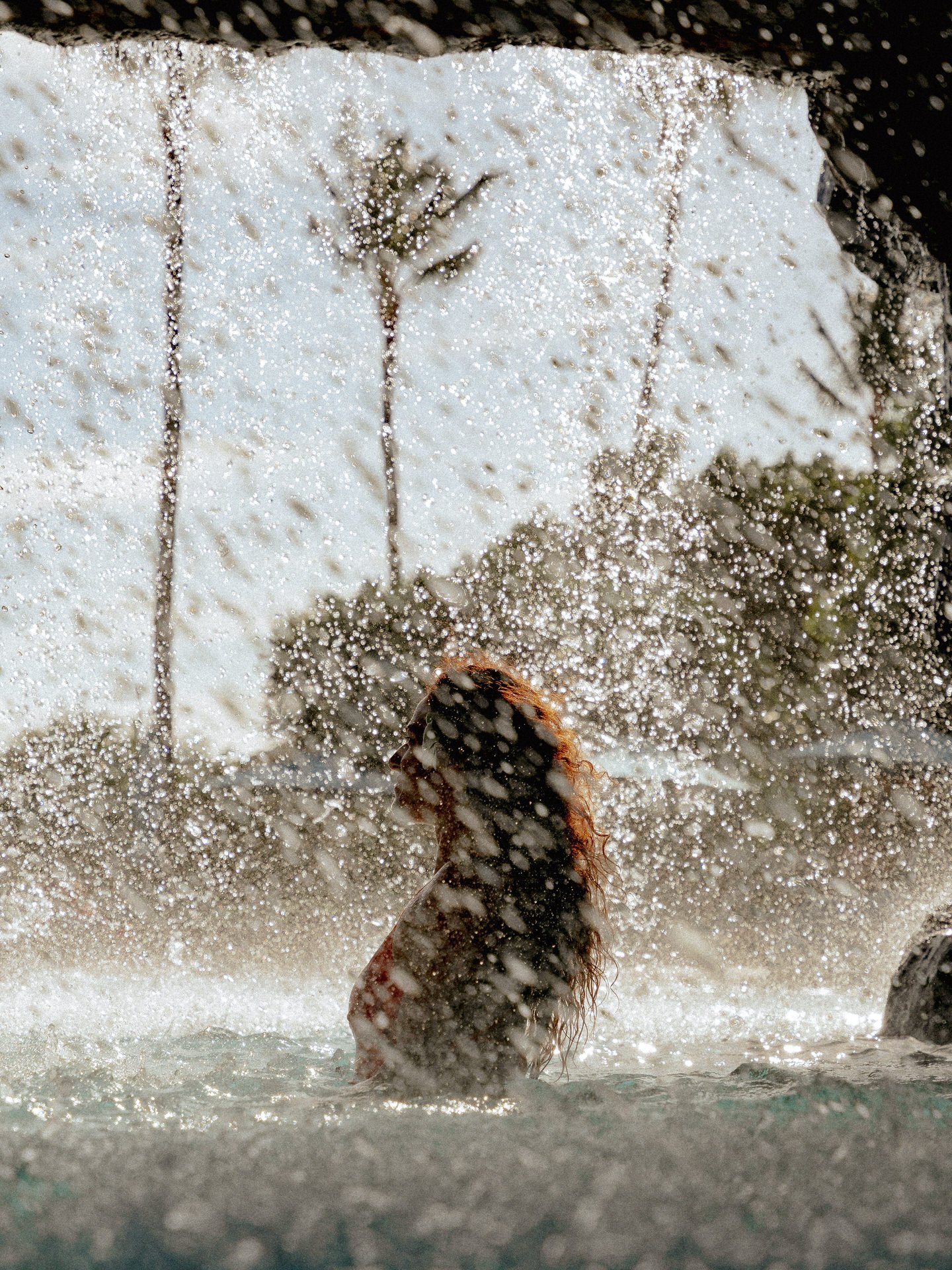 Woman with curly hair sitting in water behind a glass with water droplets, with a house and trees in the background.