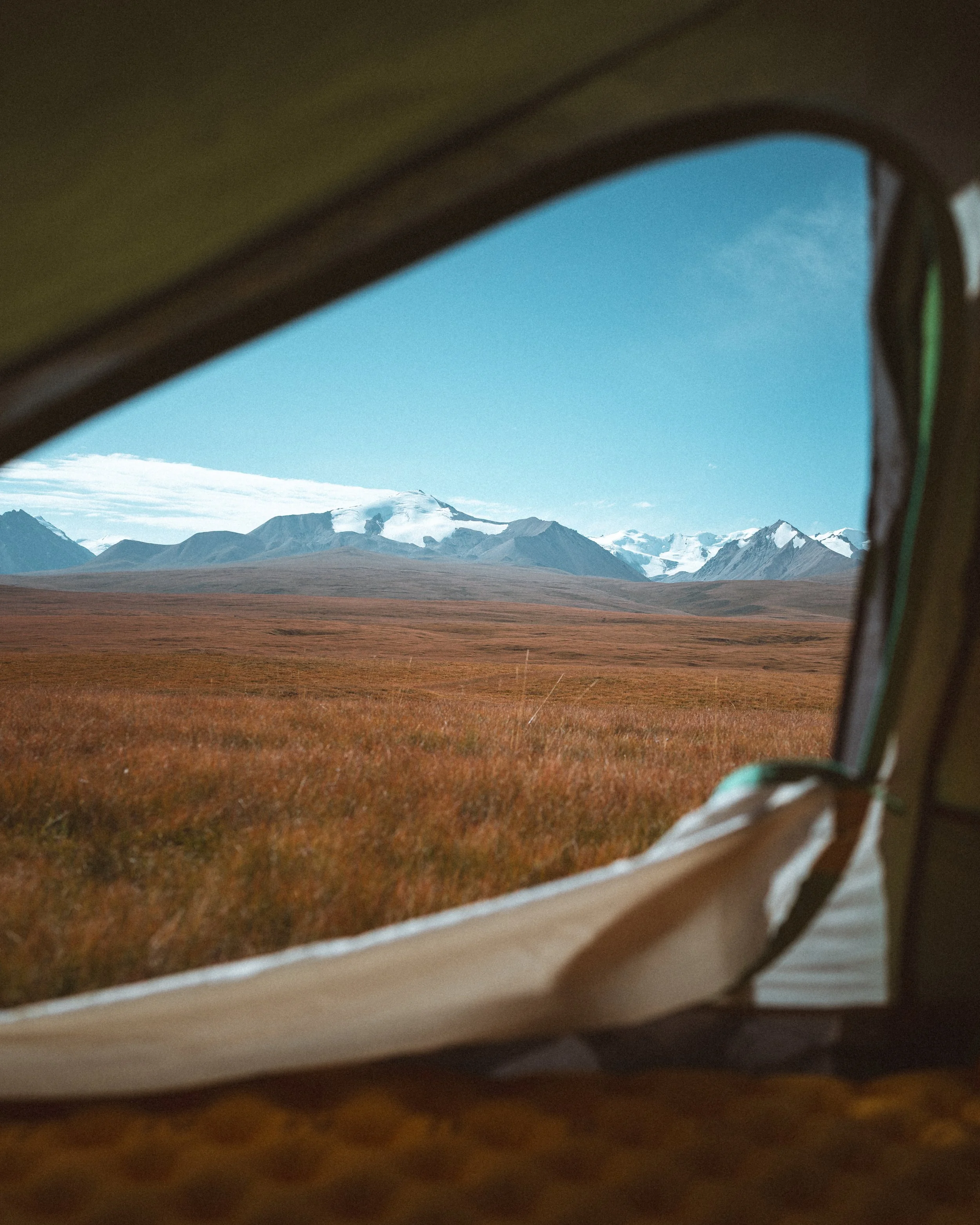 View of mountains with snow and clouds through a tent window, with a grassy plain in the foreground.