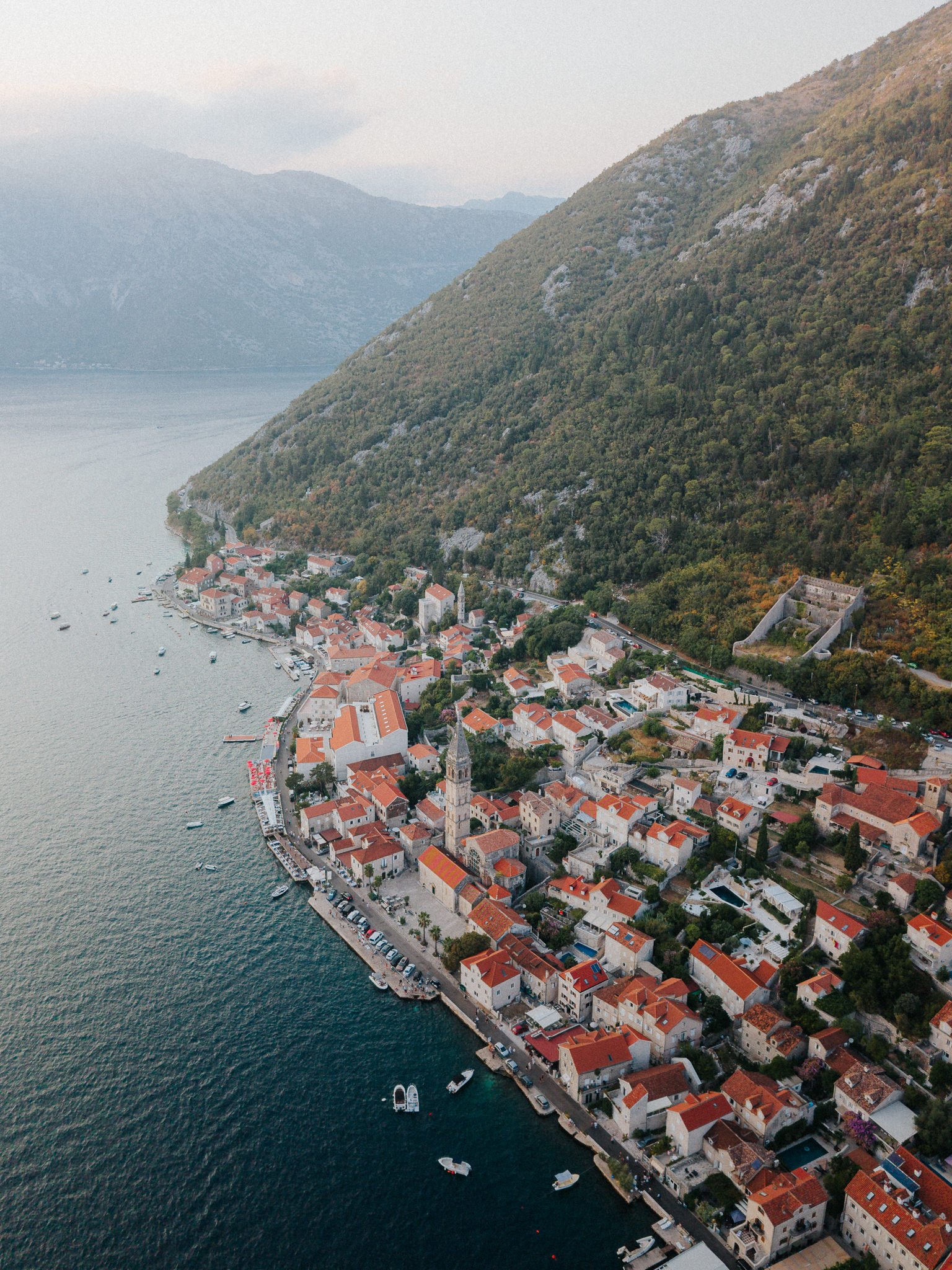Aerial view of a coastal town with red-roofed buildings, church, and boats, situated at the foot of lush green mountains with cliffs.