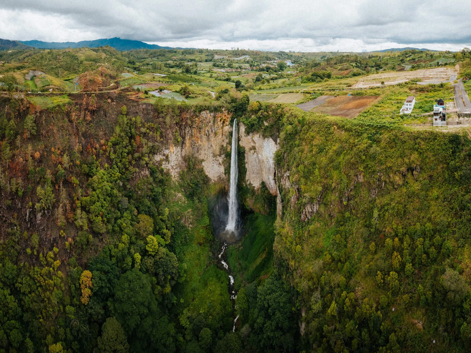 Aerial view of a tall waterfall cascading down a steep, lush green cliff surrounded by dense forest in a hilly landscape with scattered houses and farmland under a cloudy sky.