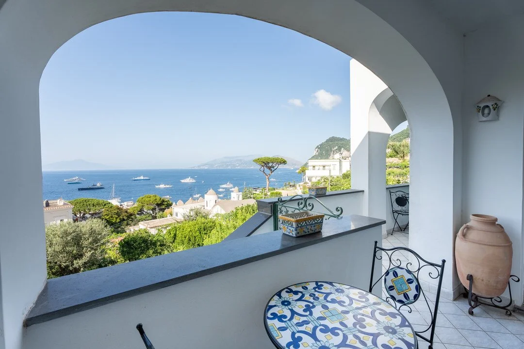 View of the ocean with boats, seen from a white balcony with decorative metal chairs and a table, in a Mediterranean setting.