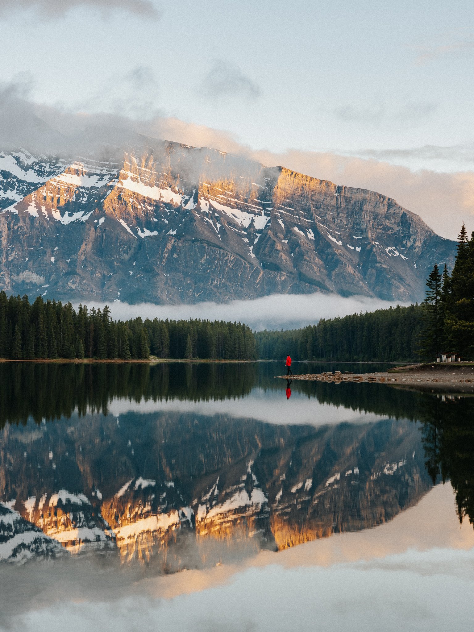 A mountain landscape with snow-capped peaks, a forest at the base, and a person in a red jacket standing by the water, with the mountain's reflection visible in the calm lake.