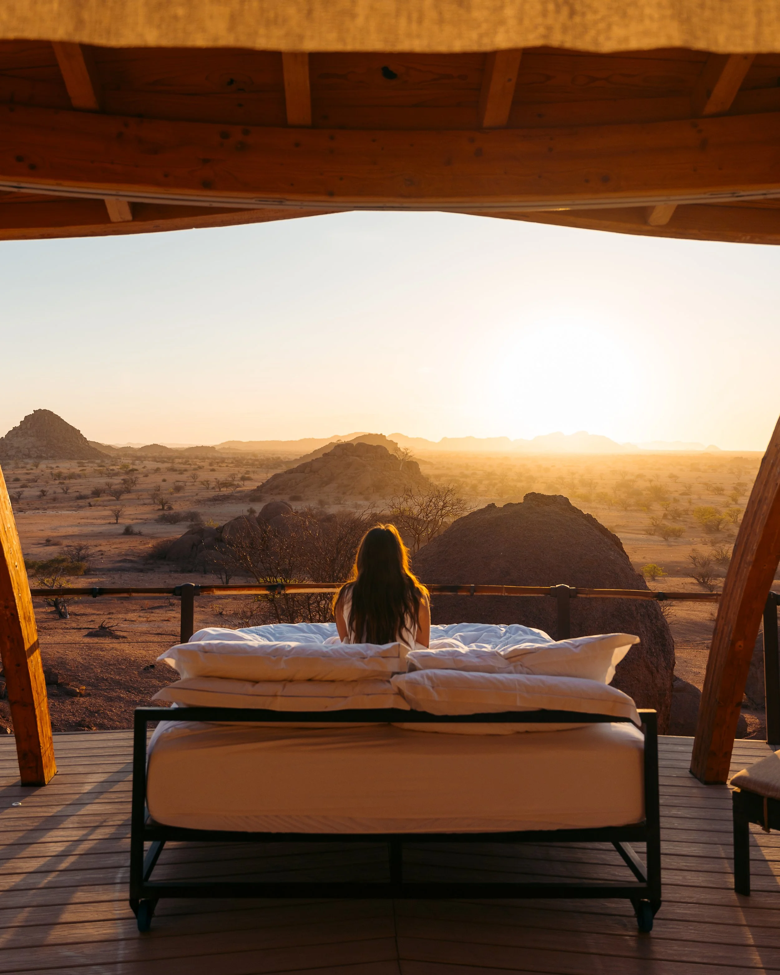 A woman with long hair sitting on a bed on a balcony, watching the sunset over a desert landscape with rocks and sparse trees.