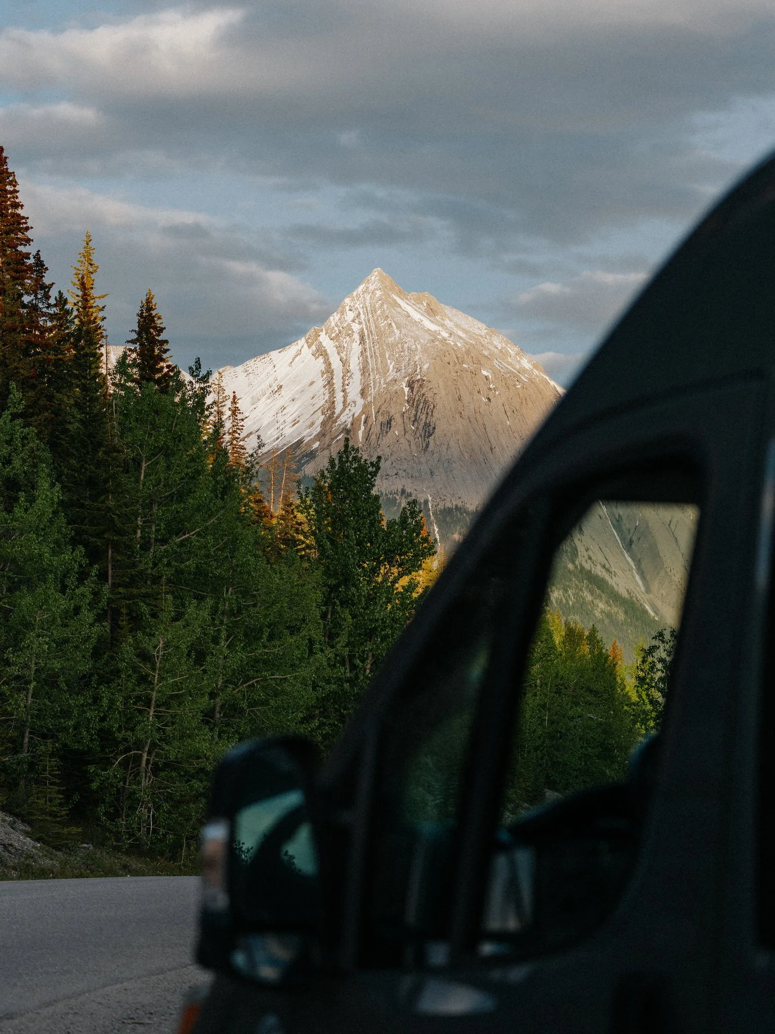 A mountain with snow on its peak and rocky slopes, viewed from inside a vehicle with a side mirror visible in the foreground, surrounded by green trees under a cloudy sky.
