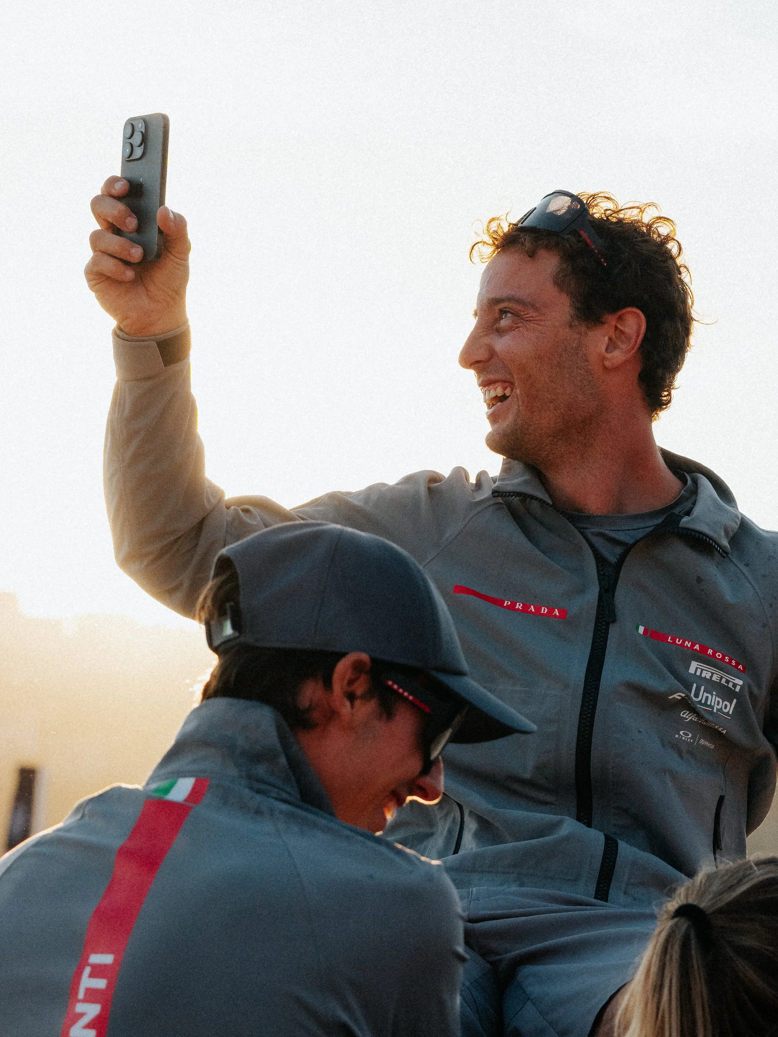 Two men in racing uniforms celebrating, with one holding a smartphone and both smiling and looking at each other, outdoors during sunset.