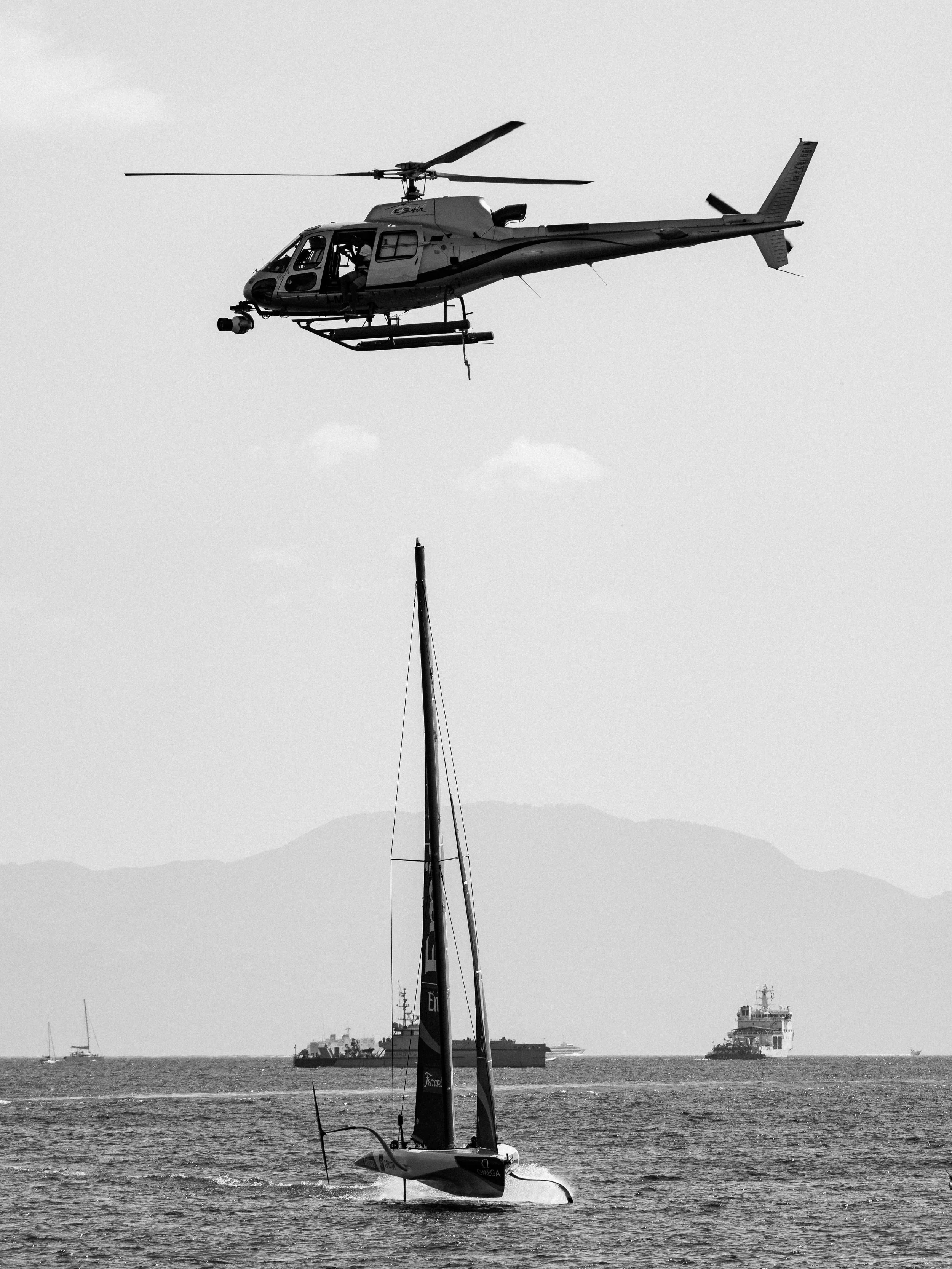 A sailboat with a hydrofoil flying above the water in a harbor, with ships and mountains in the background.