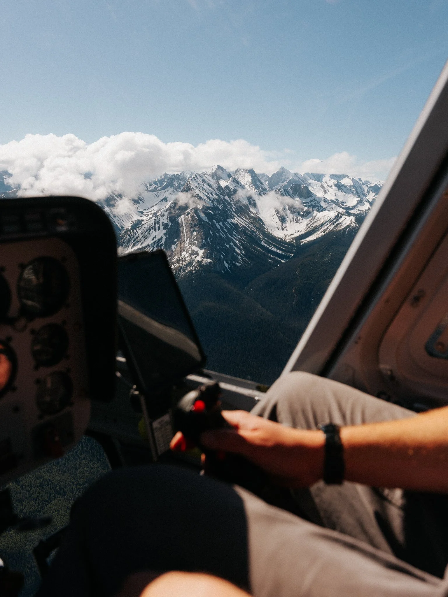Inside a helicopter cockpit with a view of snow-capped mountain peaks and clouds through the window.