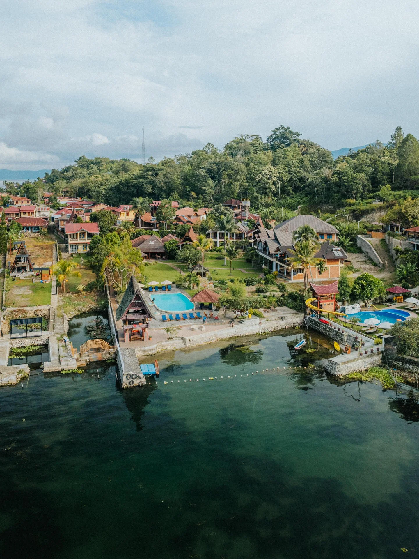 Aerial view of a resort with houses, a swimming pool, water slides, and a waterfront area surrounded by lush greenery and mountains in the background.