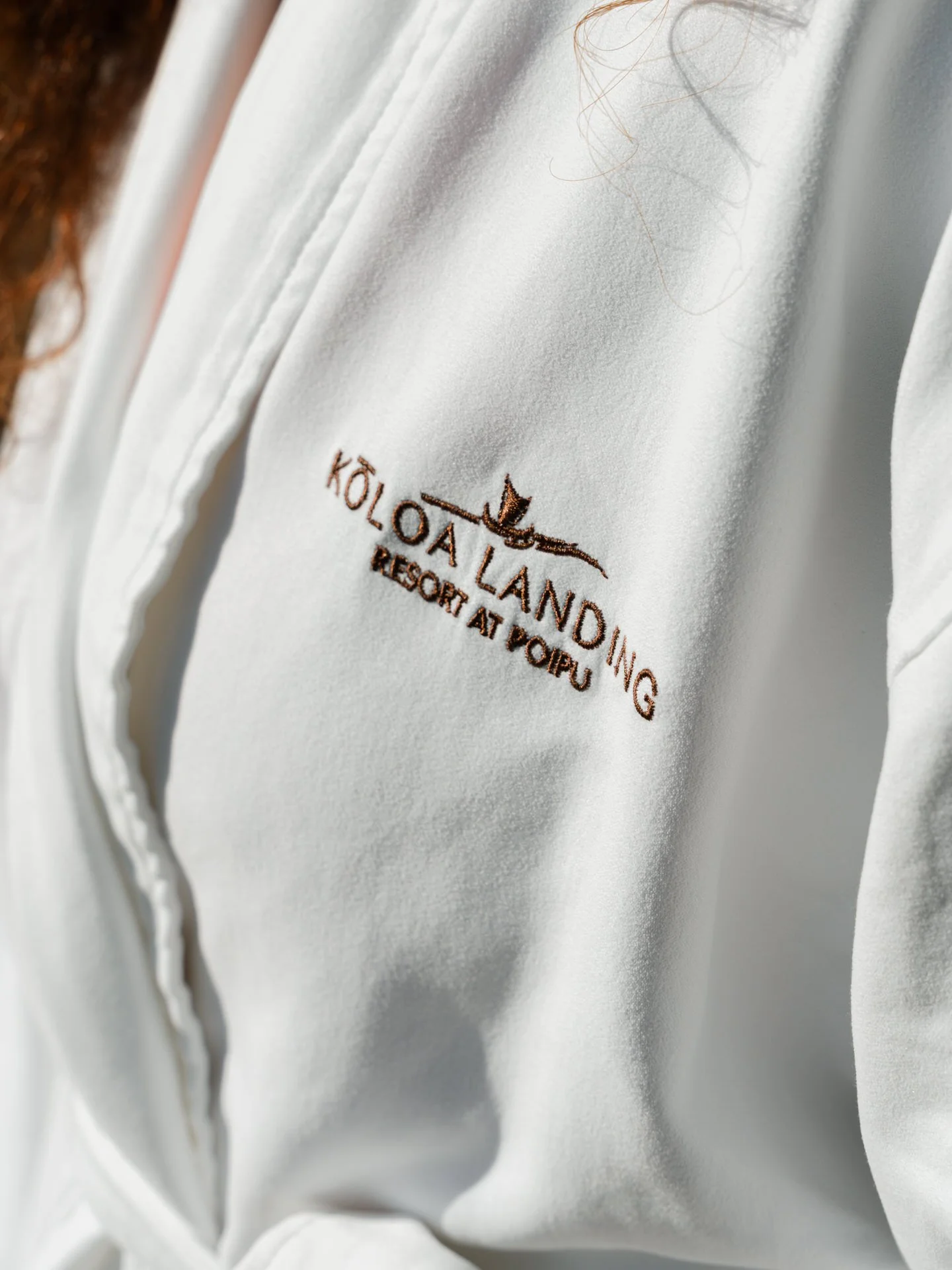 Close-up of a white jacket with embroidery of the words 'Koloa Landing Resort at Poipu' and a small logo of a bird in brown thread.