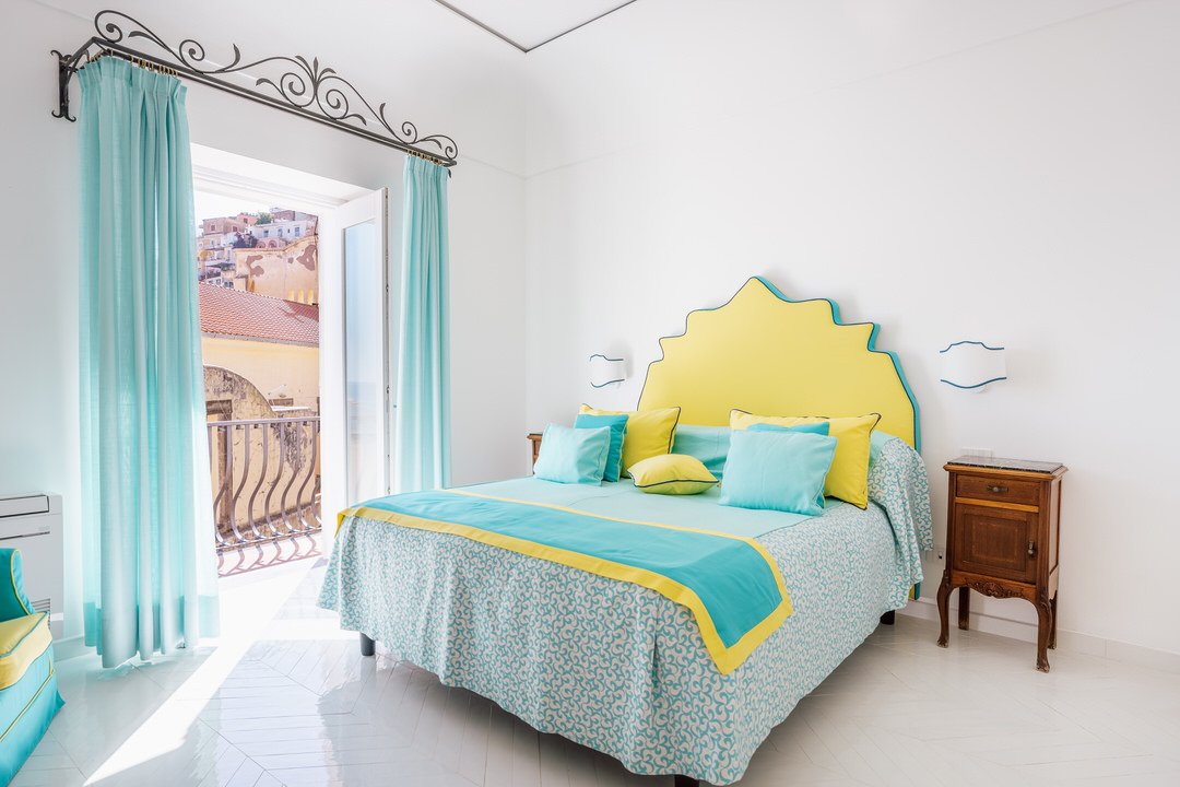 Bright bedroom with white walls, turquoise and yellow bedding, a headboard with a yellow scalloped design, wooden nightstands, light blue curtains, and a balcony door showing a Mediterranean town view.