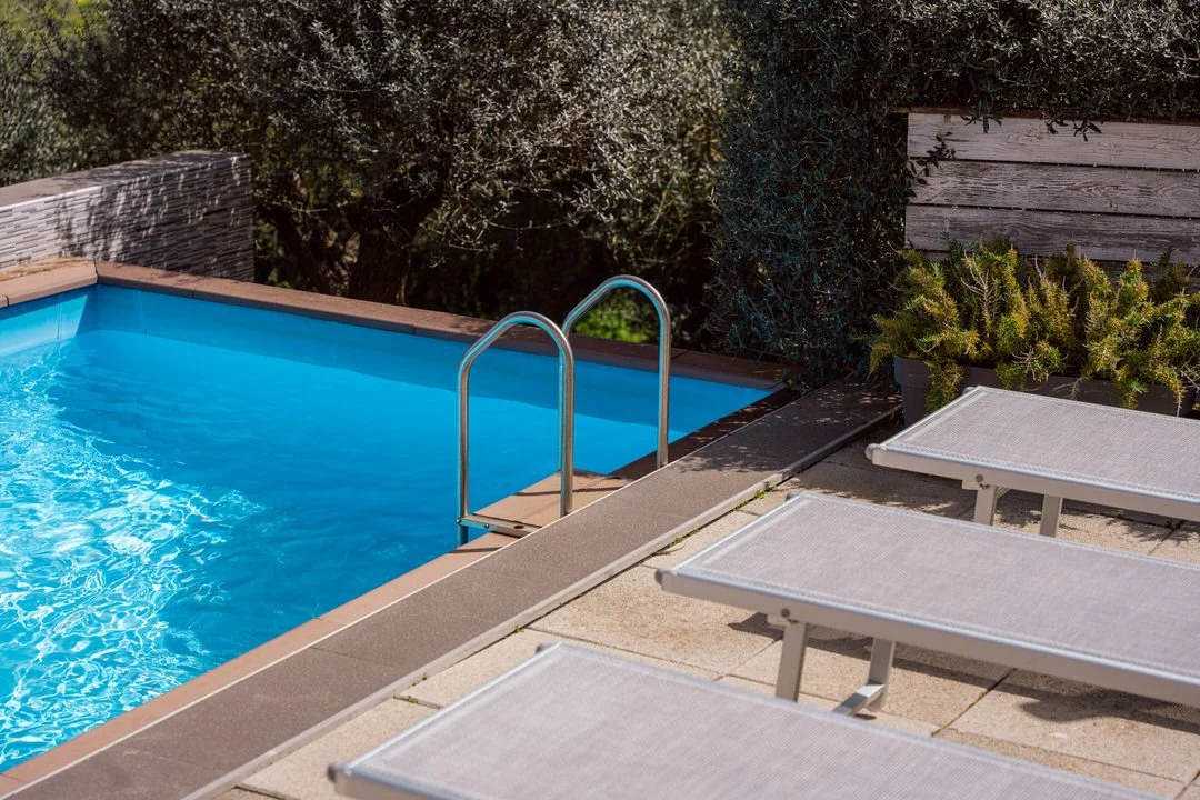 A small outdoor swimming pool with clear blue water, surrounded by a concrete deck with two lounge chairs and some greenery in the background.