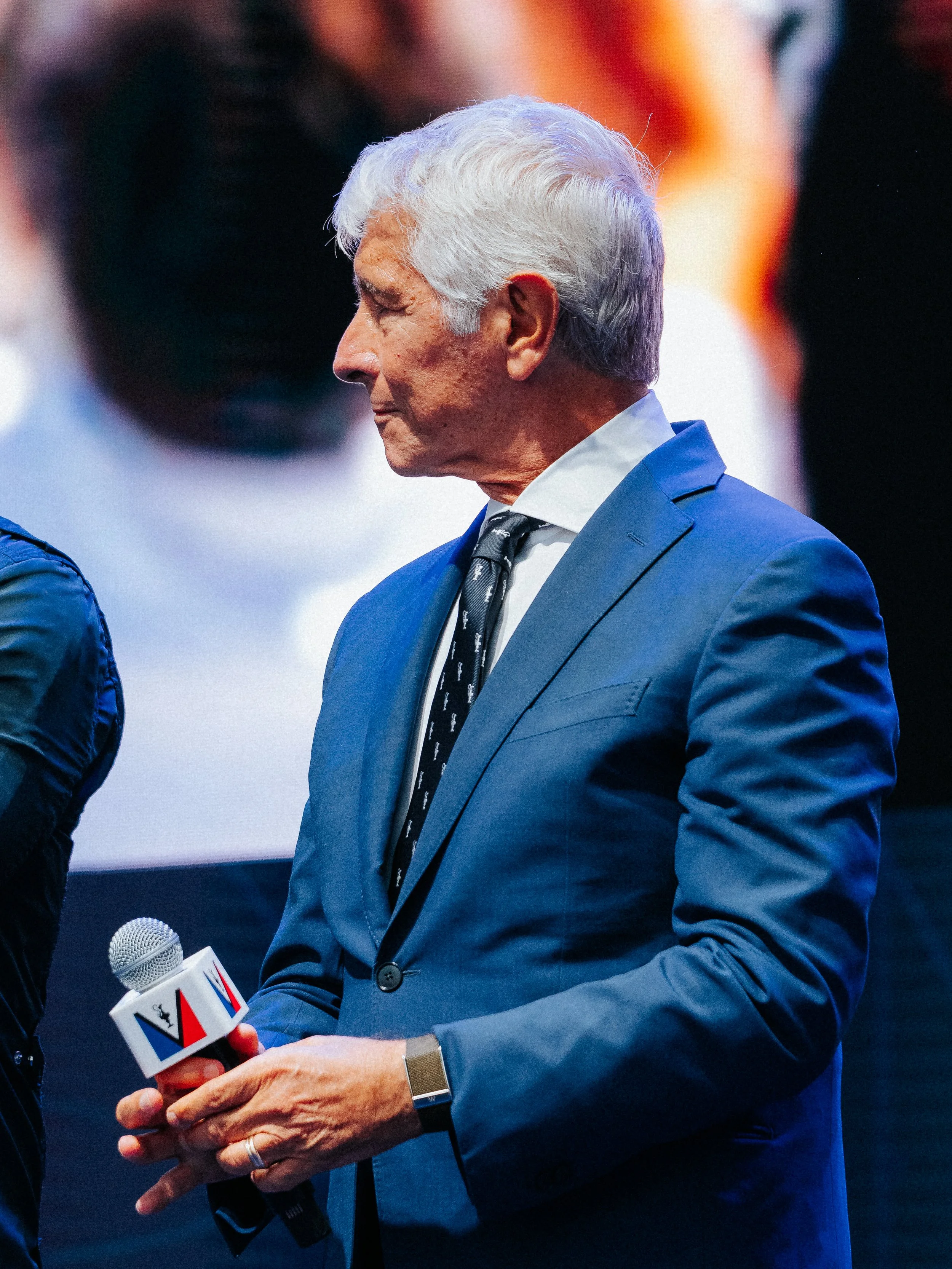 An older man in a blue suit and a patterned tie is speaking into a microphone held by someone else. The man has white hair and is wearing a smartwatch.