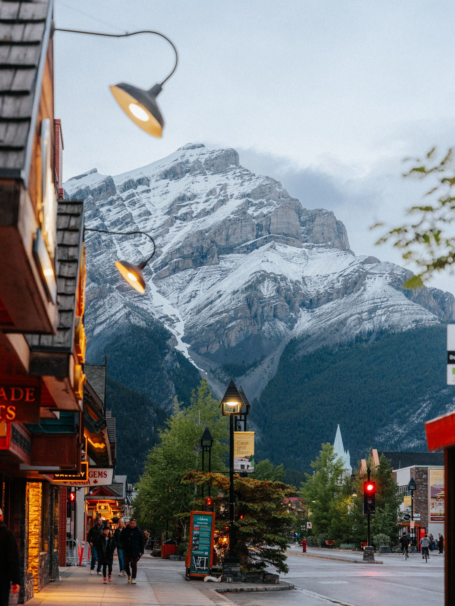 A downtown street scene with people walking, small shops and businesses on the left, street lamps, trees, and a mountain with snow in the background.