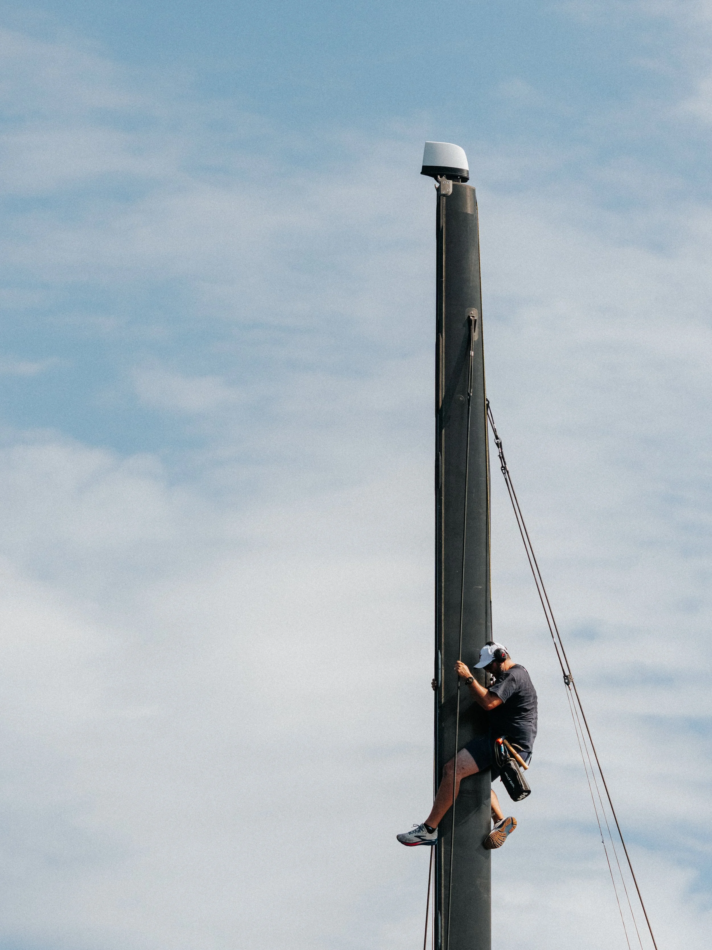 A person working on the mast of a sailboat, wearing a gray shirt, shorts, and a white cap, with a background of a light blue sky with thin clouds.