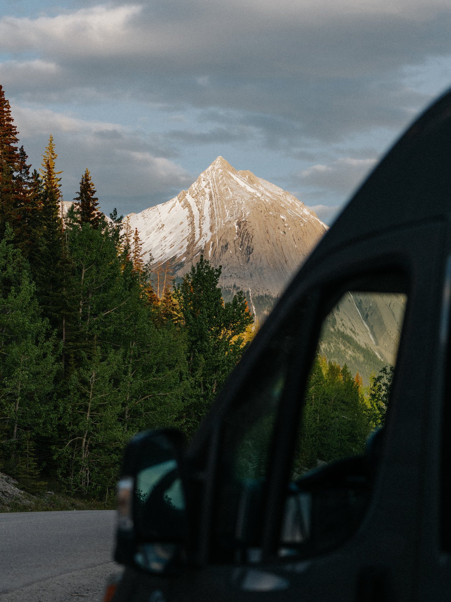 A snow-capped mountain peak seen through a side mirror of a vehicle, with trees and a cloudy sky in the background.