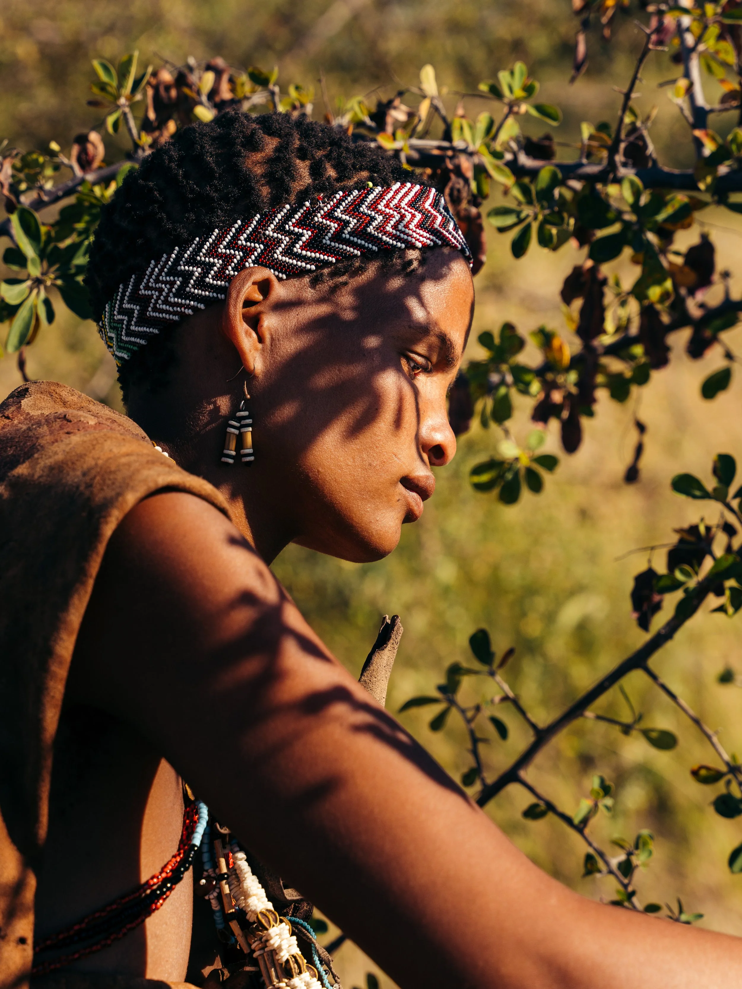Close-up of a person with curly hair wearing a beaded headband and earrings, with a beaded necklace, outdoors with greenery.